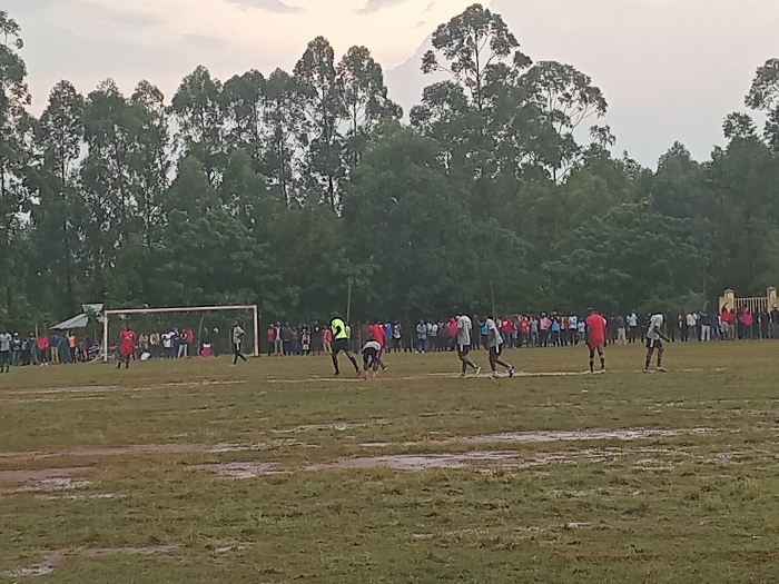 A soccer game is in progress on a muddy field. Players wearing red and light-colored jerseys are actively playing. A soccer goal is visible in the background with a crowd of spectators lining the sidelines. Tall trees dominate the background and a cloudy sky is overhead.