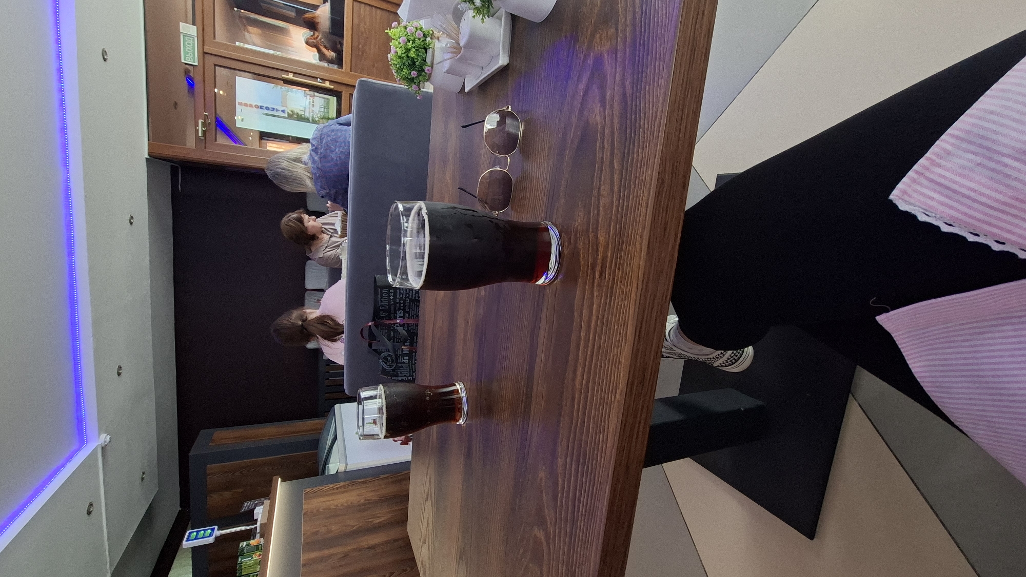 The image shows two glasses of dark beer on a wooden table in what appears to be a cafe or restaurant. In the background, several people are seated at tables. The decor includes a blue LED light strip along the ceiling and a sign that reads "ВЫХОД" (Exit) on a glass door. The location is identified as Myadzyel, Belarus. The foreground shows a person's legs, wearing black leggings and white sneakers, with a pink striped fabric item. Sunglasses are placed on the table next to one of the beer glasses.