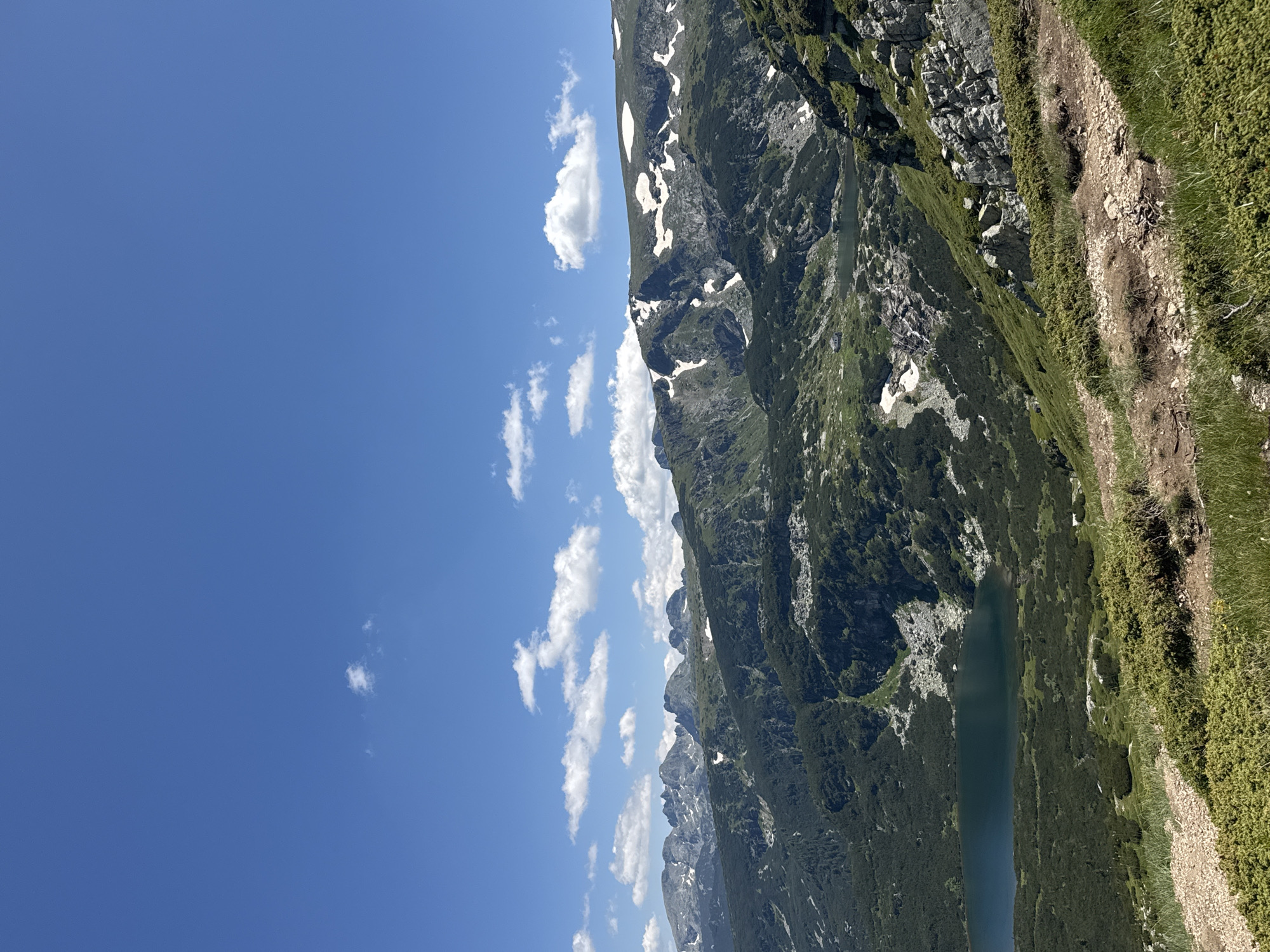 This is a scenic, eye-level, wide-angle photograph taken from a mountainous hiking trail. The sky is a clear, bright blue with scattered white cumulus clouds. In the middle ground, a dark blue, serene lake is nestled among rolling green mountains covered with dense, dark green vegetation, possibly pine trees. Patches of snow are visible on the higher slopes of the mountains, suggesting a high altitude. The foreground consists of a rough, dirt hiking path with sparse green grass and low-lying, dense green bushes. The overall scene depicts a natural, mountainous landscape, likely in the Rila Mountains near Panichishte, Bulgaria, a popular tourist destination known for its glacial lakes and hiking trails. There are no people or animals visible in the image.