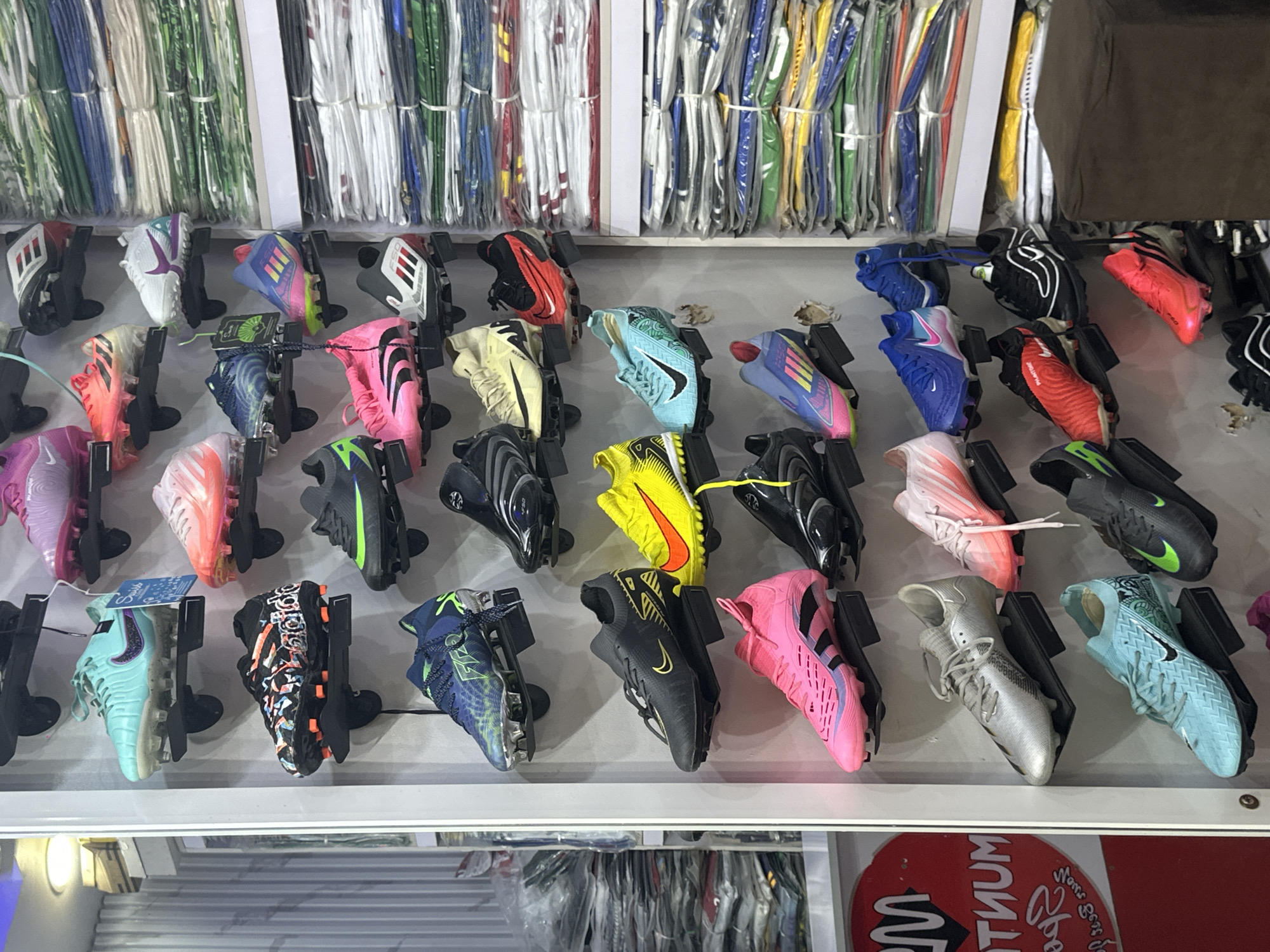 The image depicts a retail display of athletic footwear, specifically football boots, arranged on tiered shelving units against a light-colored wall. The boots are predominantly in bright, vibrant colors such as turquoise, pink, yellow, red, and blue, with some featuring intricate patterns and branding. Several pairs are visibly new, with tags still attached.
To the right of the footwear display, stacks of folded clothing items, likely sports apparel like jerseys or tracksuits, are neatly arranged on shelves. These items are in various colors, including green, blue, white, and red, and are wrapped in plastic.
On the far left, a partial view of a red circular sign with white lettering is visible. The lettering appears to be in a stylized font and reads "M NTUM" and some other illegible text below. This sign suggests a commercial establishment.
The overall environment suggests a retail store specializing in sportswear. While no people are directly visible in the frame, the arrangement of merchandise implies a business setting. The image does not contain any specific visual cues to definitively place the location in Jos, Nigeria, or any other specific city. However, the presence of football boots and sports apparel is common in many retail outlets globally.