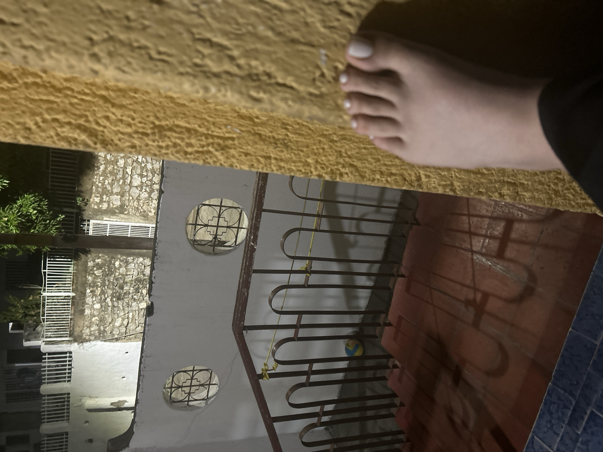 A close-up shot focuses on a person's foot with white nail polish on a rough, yellow surface. In the background, through a railing, is a view of a building. The building features decorative round windows and a wall with stone or brickwork. A ball is visible on the ground level of the building. The scene suggests a casual, possibly nighttime or low-light setting. There is no verifiable evidence in the image to support that it was taken in San Juan de Los Morros, Venezuela.
