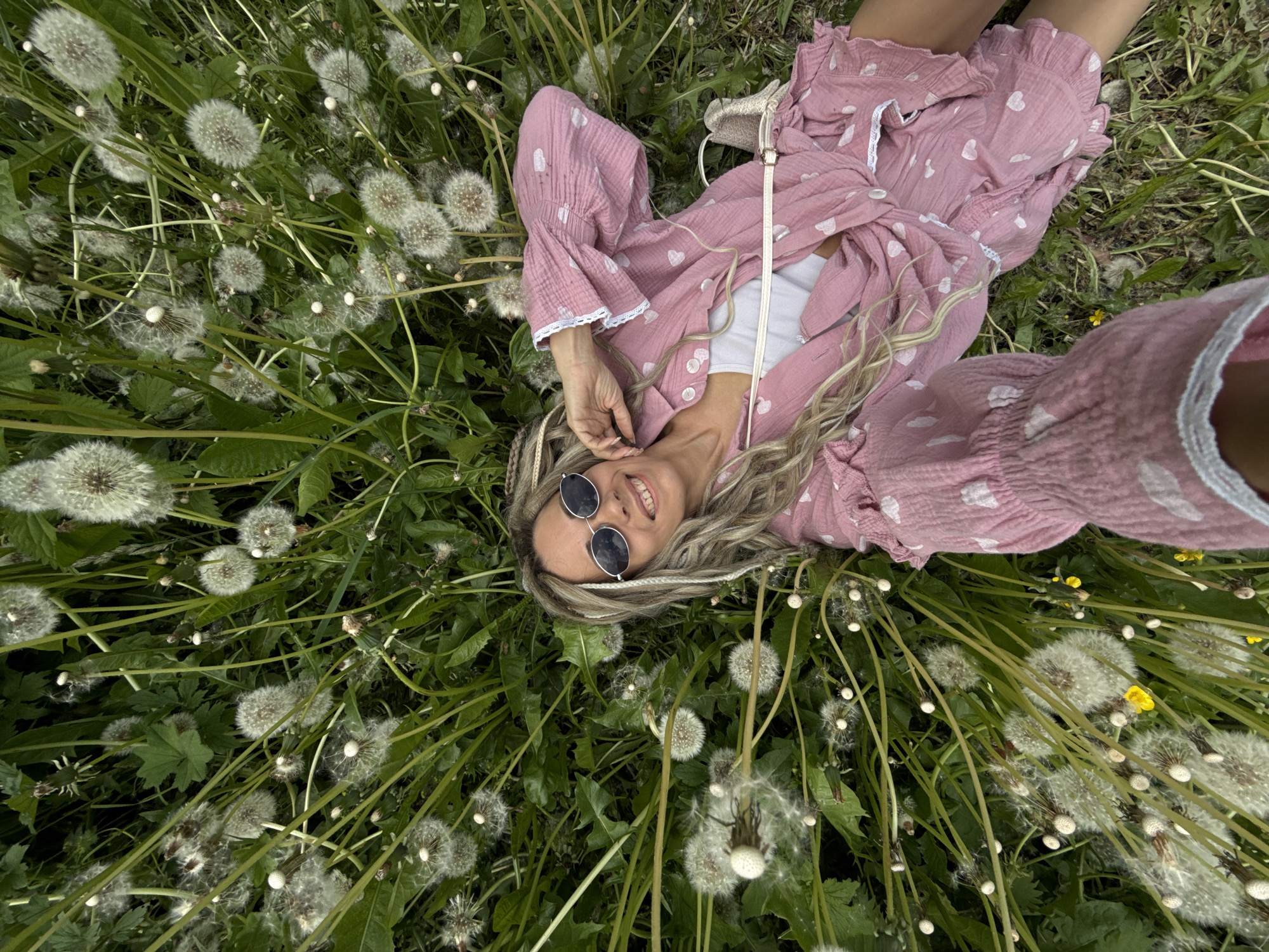 A woman is seen lying on her back in a vibrant green field filled with numerous dandelions in their mature, white seed head stage. She is positioned with her head towards the bottom of the frame, looking up and smiling broadly at the camera. Her blonde hair, which features some braided or dreaded sections, frames her face. She is wearing round, dark-lensed sunglasses. Her attire consists of a pink, long-sleeved top or dress with a ruffled texture and white heart patterns, featuring white lace trim on the cuffs and collar. A white top is visible underneath. A light-colored strap, possibly from a bag, runs across her chest. The field is densely packed with tall green grass and dandelion plants, creating a lush, natural setting. The lighting suggests it is daytime, likely sunny given the use of sunglasses. The overall mood conveyed is one of relaxation and enjoyment. No visible text is present in the image. The location context provided is Rusanovo, Russia.