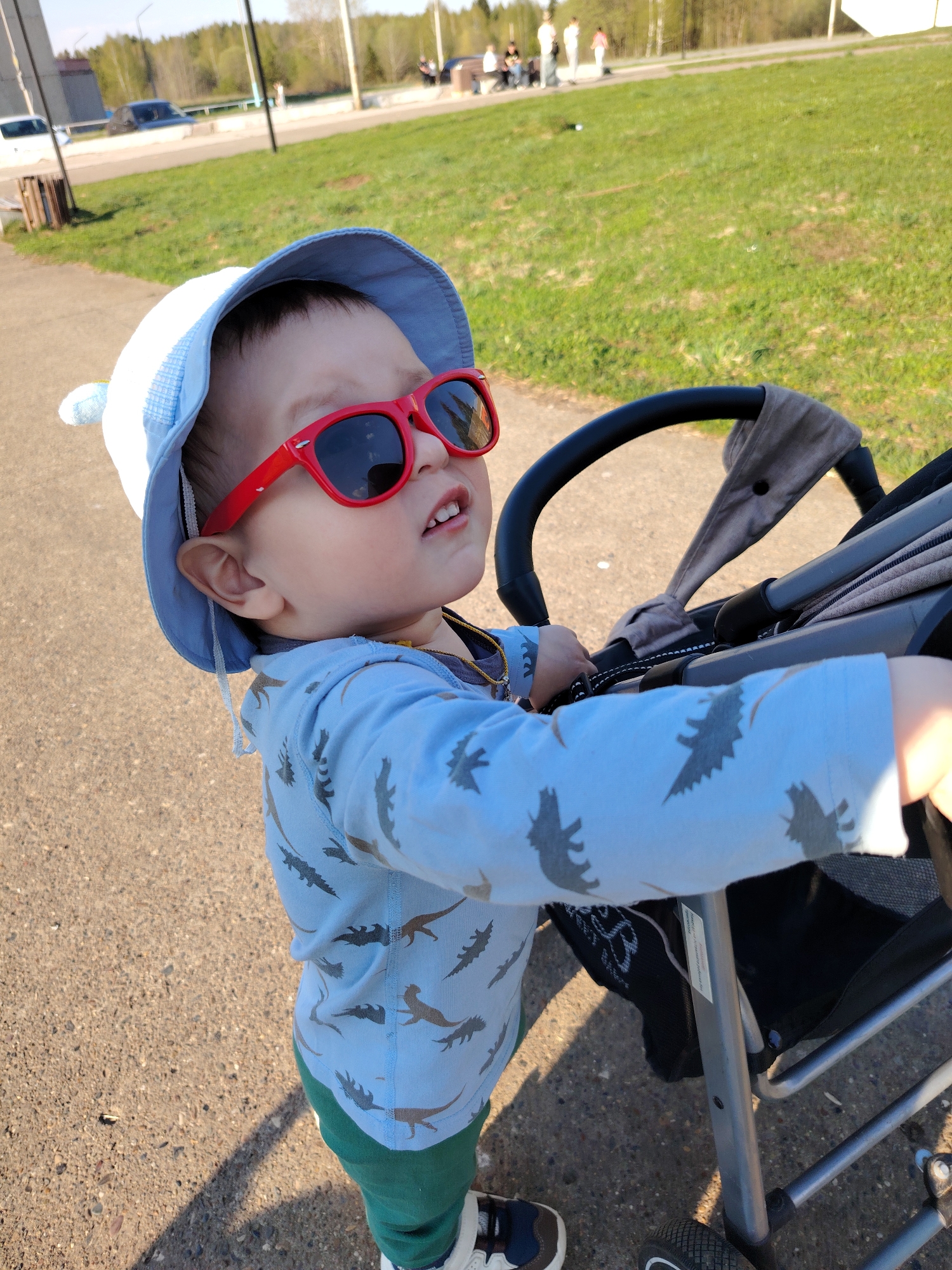 In the city of Марковский, Russia, a child wearing red sunglasses and a blue hat stands next to a stroller on a paved path. The child is looking upwards. The path is bordered by grass. In the background, there are trees and other people.