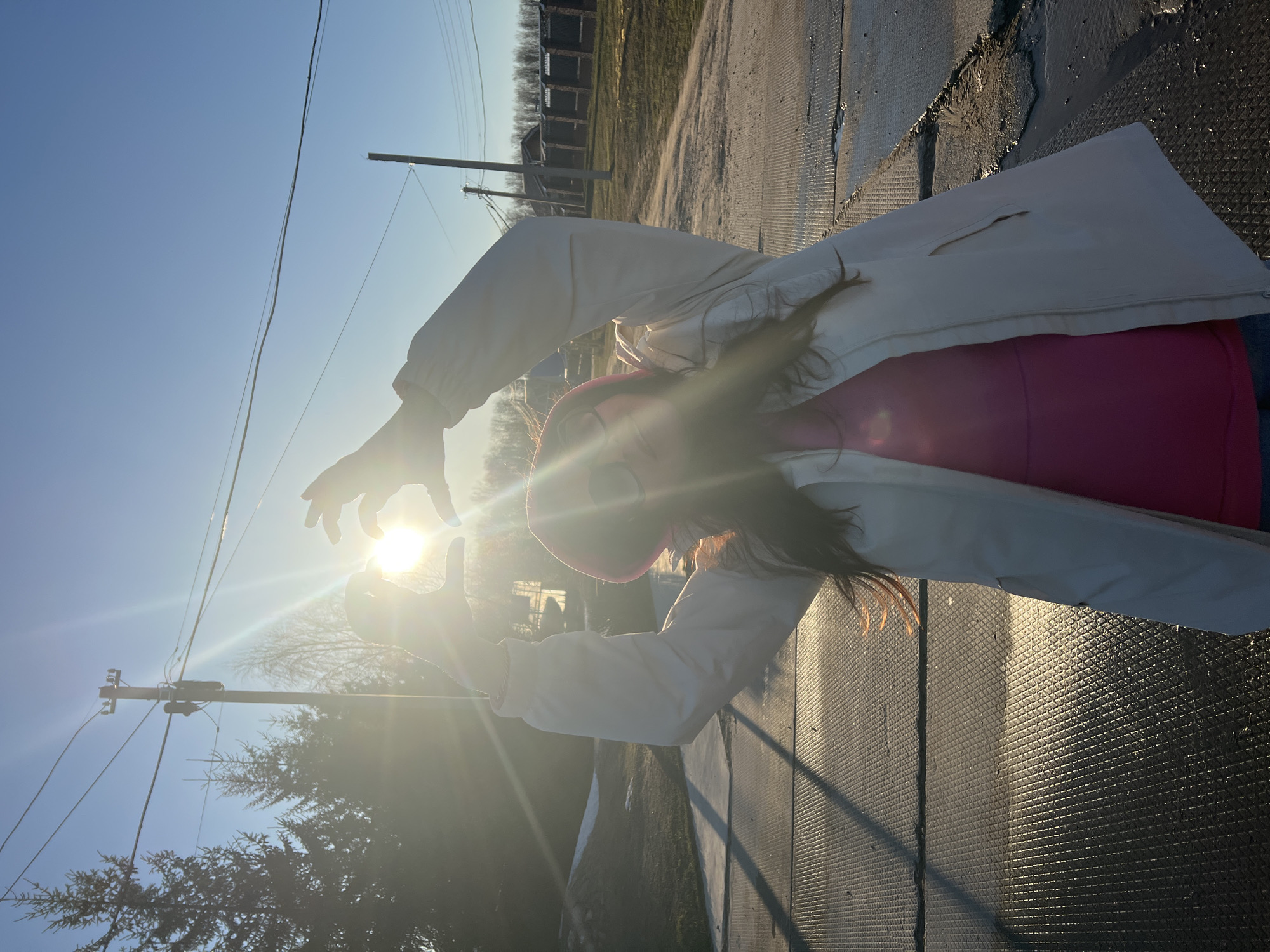 A young woman in a pink shirt and white jacket poses with her hands near the sun, obscuring her face. The background features a blue sky, power lines, trees, and a building. The location is outdoors, seemingly near a road or pathway. There is no indication this location is specifically in the city of Noviy, Russia.