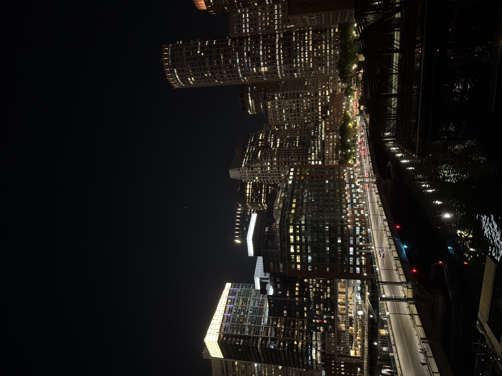 The image depicts the Boston skyline at night, seen from a high vantage point. Several illuminated skyscrapers line the waterfront. A bridge spans a dark body of water, reflecting the city lights. The cityscape is characterized by the contrast between the darkness of the night sky and the bright lights emanating from the buildings. Traffic is visible moving along the bridge.