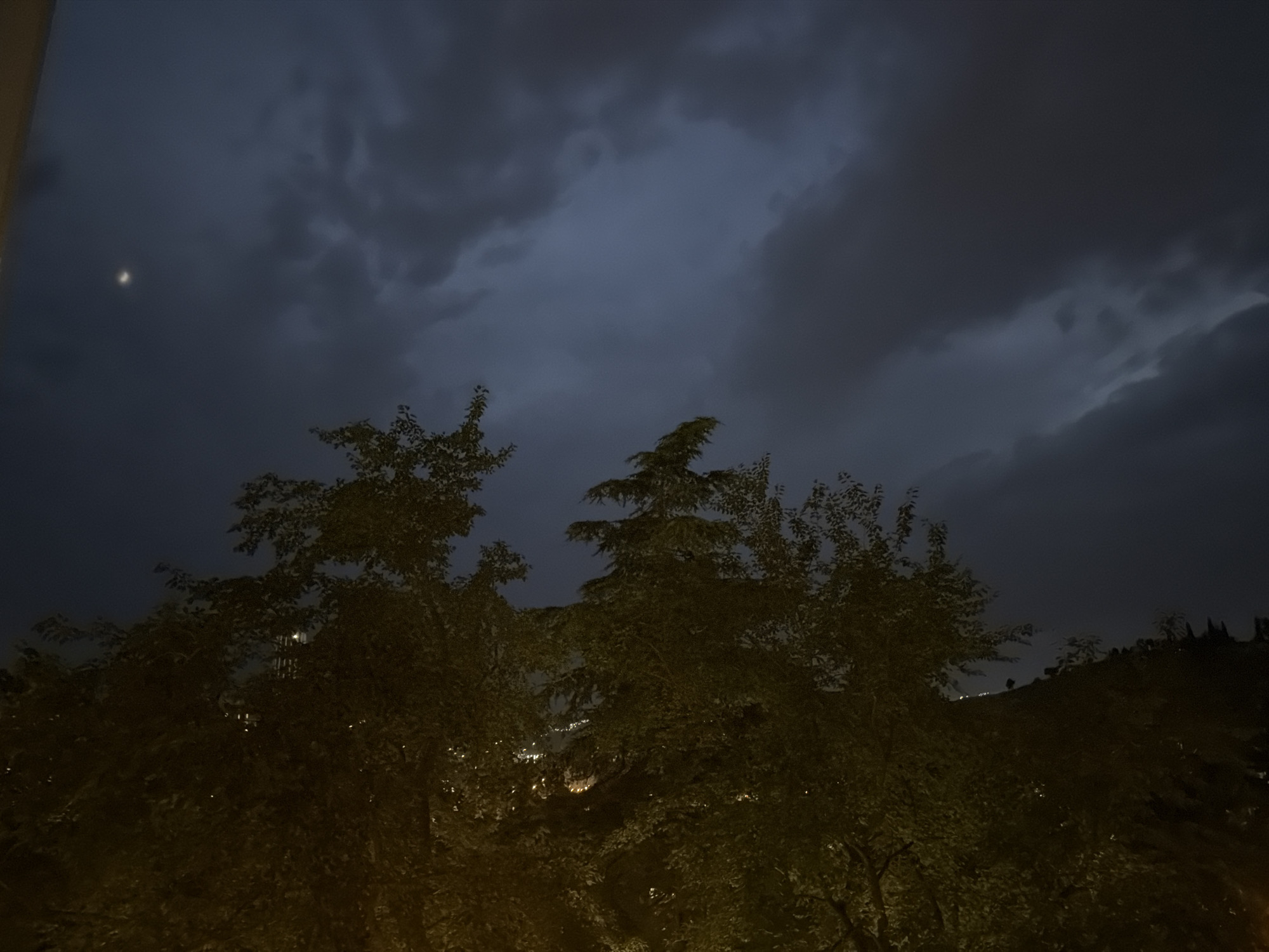 The image shows a night scene with a dark, cloudy sky and trees in the foreground. There is a faint crescent moon visible in the upper left quadrant of the sky. The trees obscure a distant view of city lights, suggesting an urban or suburban environment. The overall impression is one of a quiet, overcast evening. The location is identified as Tbilisi, Georgia, though no specific landmarks are discernible from the image itself.