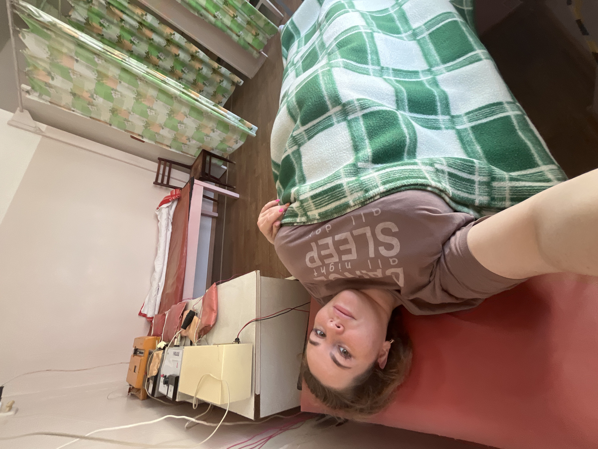 A woman lies under a green and white plaid blanket on a red surface, presumably a bed or examination table. The woman has light skin, light-colored eyes, and shoulder-length brown hair. She is wearing a light brown t-shirt with the word "SLEEP" printed on it. A series of medical devices and cords are visible on a white surface. The scene appears to be located in a medical or therapeutic setting. In the background is a green and white curtain. Based on context cues such as medical devices, the photo appears to be taken at a health facility in Chaikovsky, Russia.