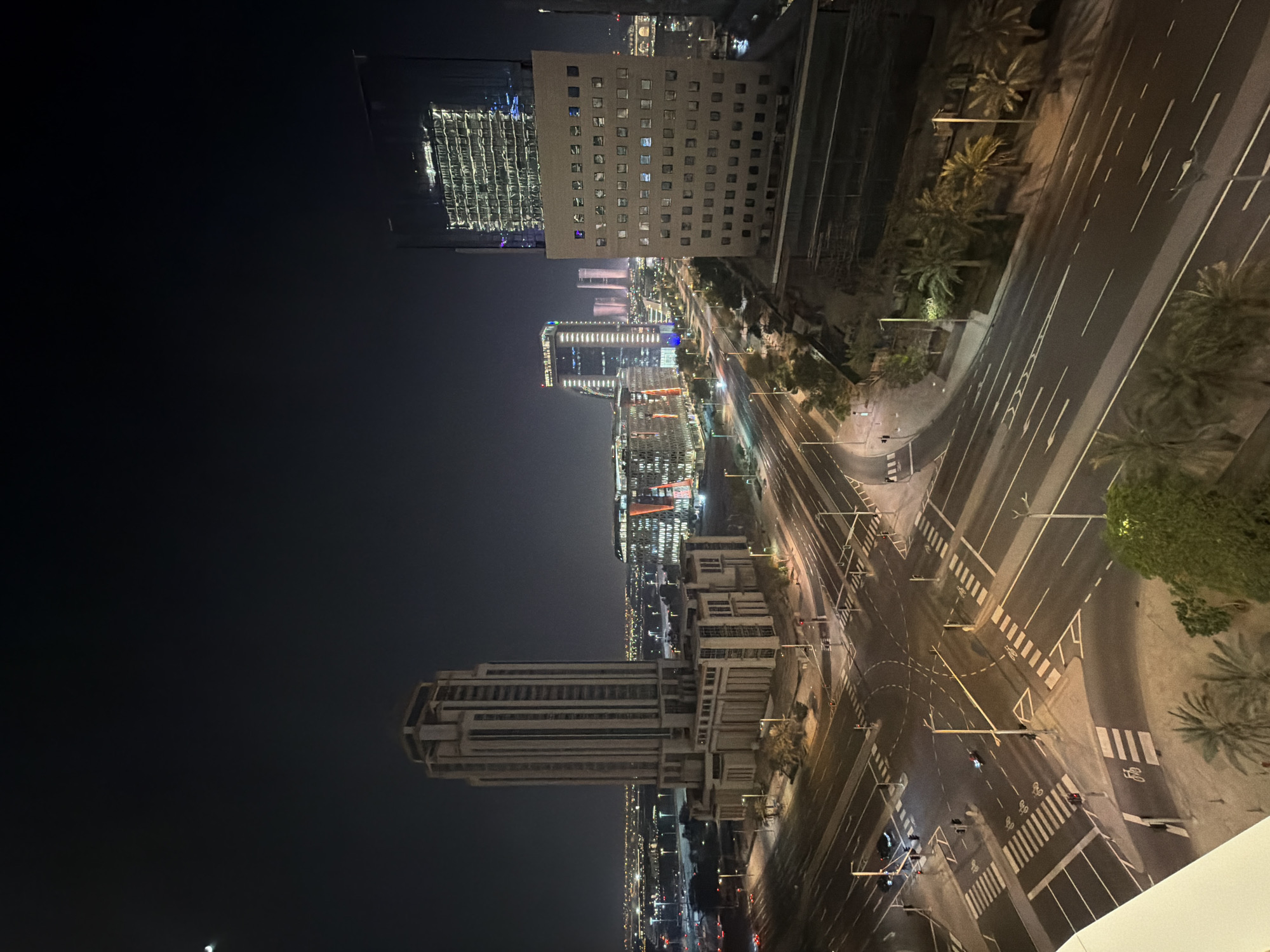 The image is an aerial nighttime view of a city intersection with several modern high-rise buildings. The sky is dark, and the city lights are illuminated. The most prominent building on the left is a tall, light-colored, rectangular skyscraper with a stepped top. In the center of the image, there are several other modern buildings, some with glass facades that reflect the city lights. One building on the right has a facade covered in numerous small square windows. The intersection below is well-lit, with multiple lanes of asphalt roads marked with white lines and pedestrian crosswalks. Traffic lights are visible at the intersection. There are a few cars on the roads, and streetlights illuminate the scene. Palm trees are sparsely scattered around the intersection and the buildings. The overall impression is a contemporary urban landscape at night.