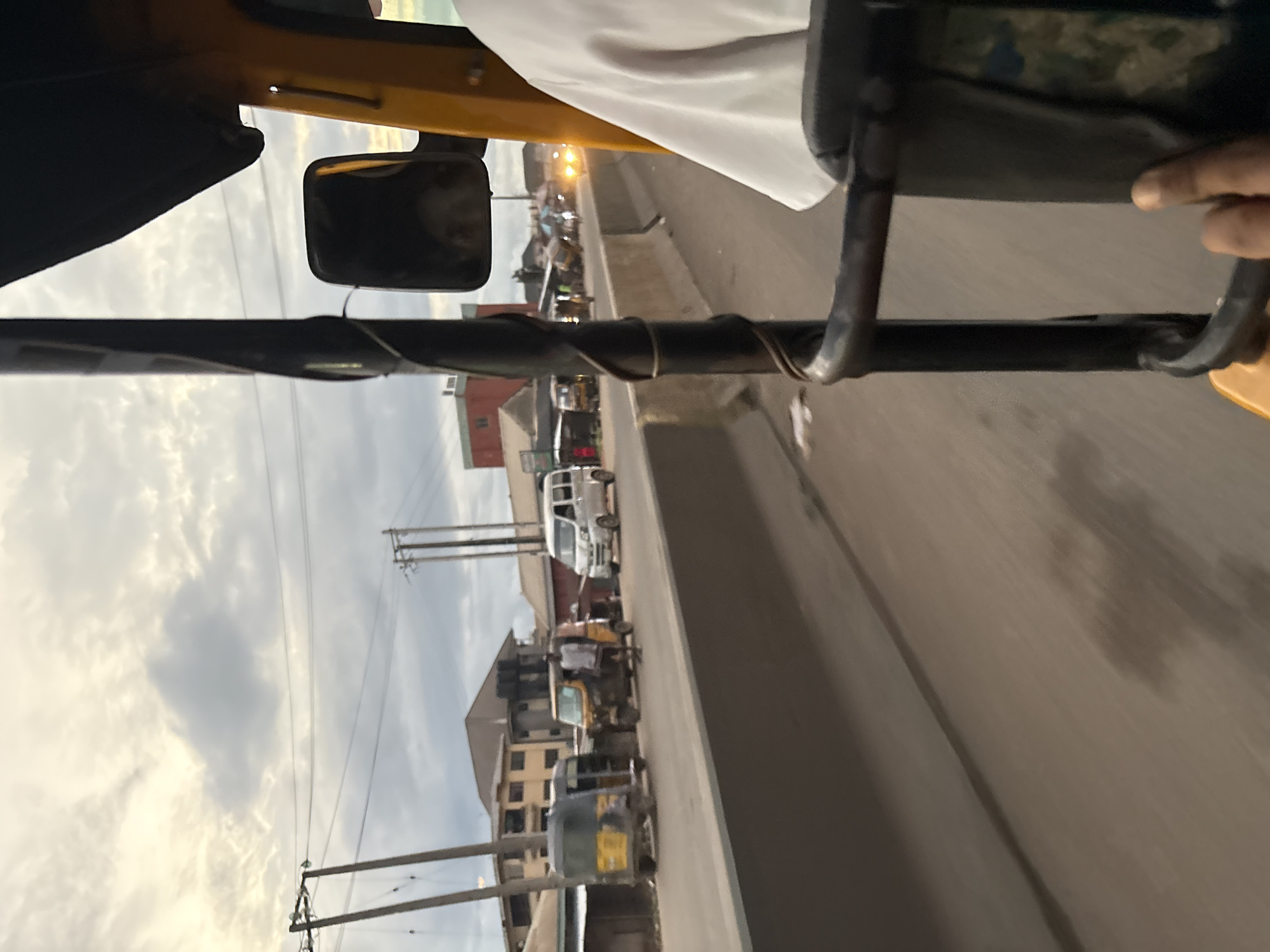 The image captures a street scene in Onitsha, Nigeria, viewed from the perspective of someone inside a yellow auto-rickshaw. The sky is overcast with some hints of light peeking through the clouds. In the foreground, a black metal bar from the auto-rickshaw's frame runs vertically through the image, with a spiral of wire wrapped around it. To the right, a person wearing a white shirt is visible, with their arm extending out of the frame.
The road is a busy thoroughfare with a concrete barrier separating lanes. Several vehicles are present on the road, including other auto-rickshaws, a white van, and a few other cars. Some of the auto-rickshaws appear to be yellow and black. On the left side of the road, buildings line the street, with some appearing to be shops or residential structures. Utility poles with wires are also visible along the roadside. The overall impression is that of a typical urban environment in Nigeria.