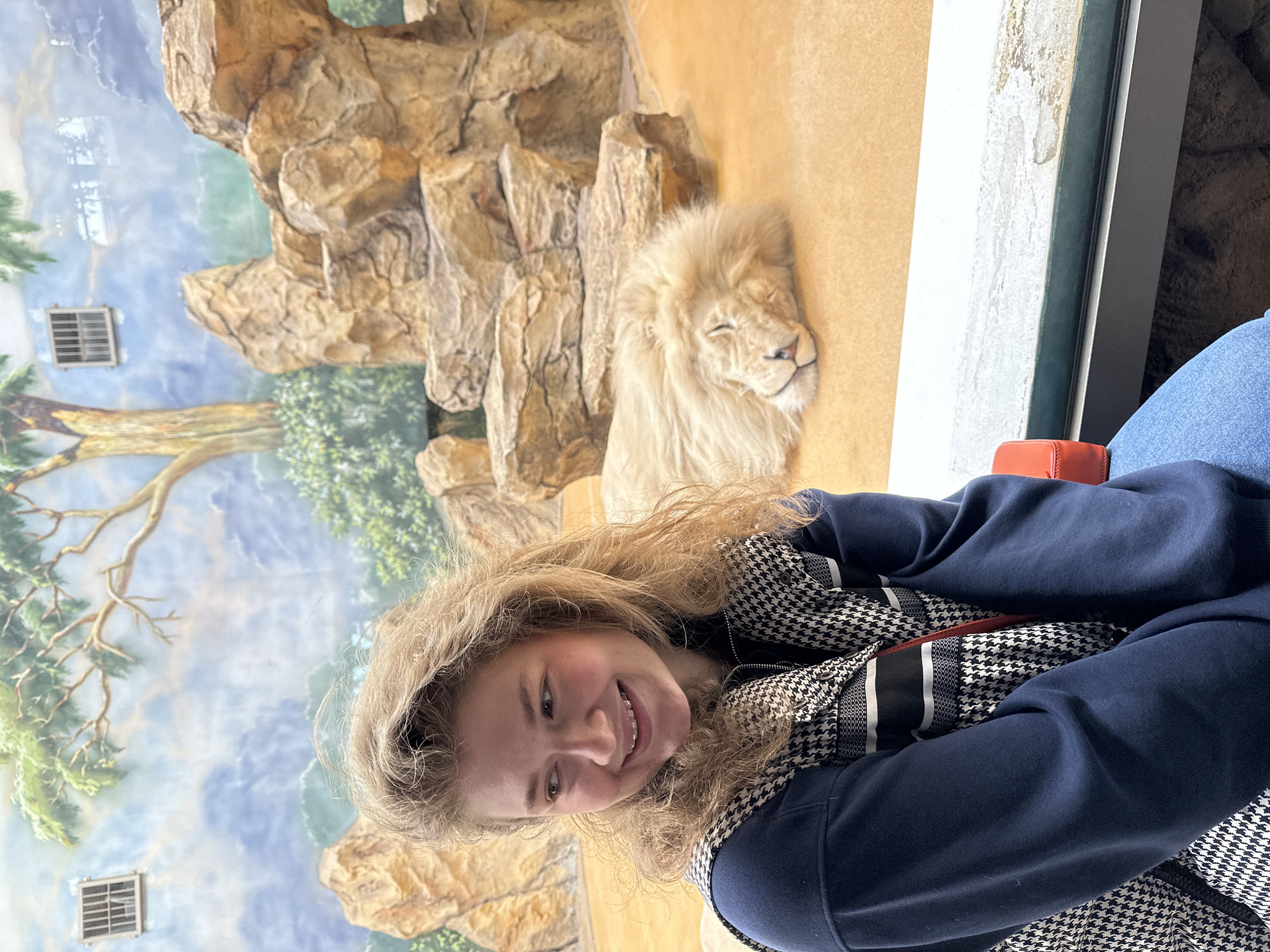 A young woman with fair skin, blonde hair, and braces is smiling in front of an enclosure. A lion is inside the enclosure. This photo was taken at the Kazan Zoo in Kazan, Russia. The lion is lying down with its eyes closed. The enclosure appears to be indoors with painted or artificial rocks and a tree scene in the background. The woman is wearing a dark blue hooded sweater and a black and white houndstooth patterned jacket or scarf. A reddish-orange object is visible near her.