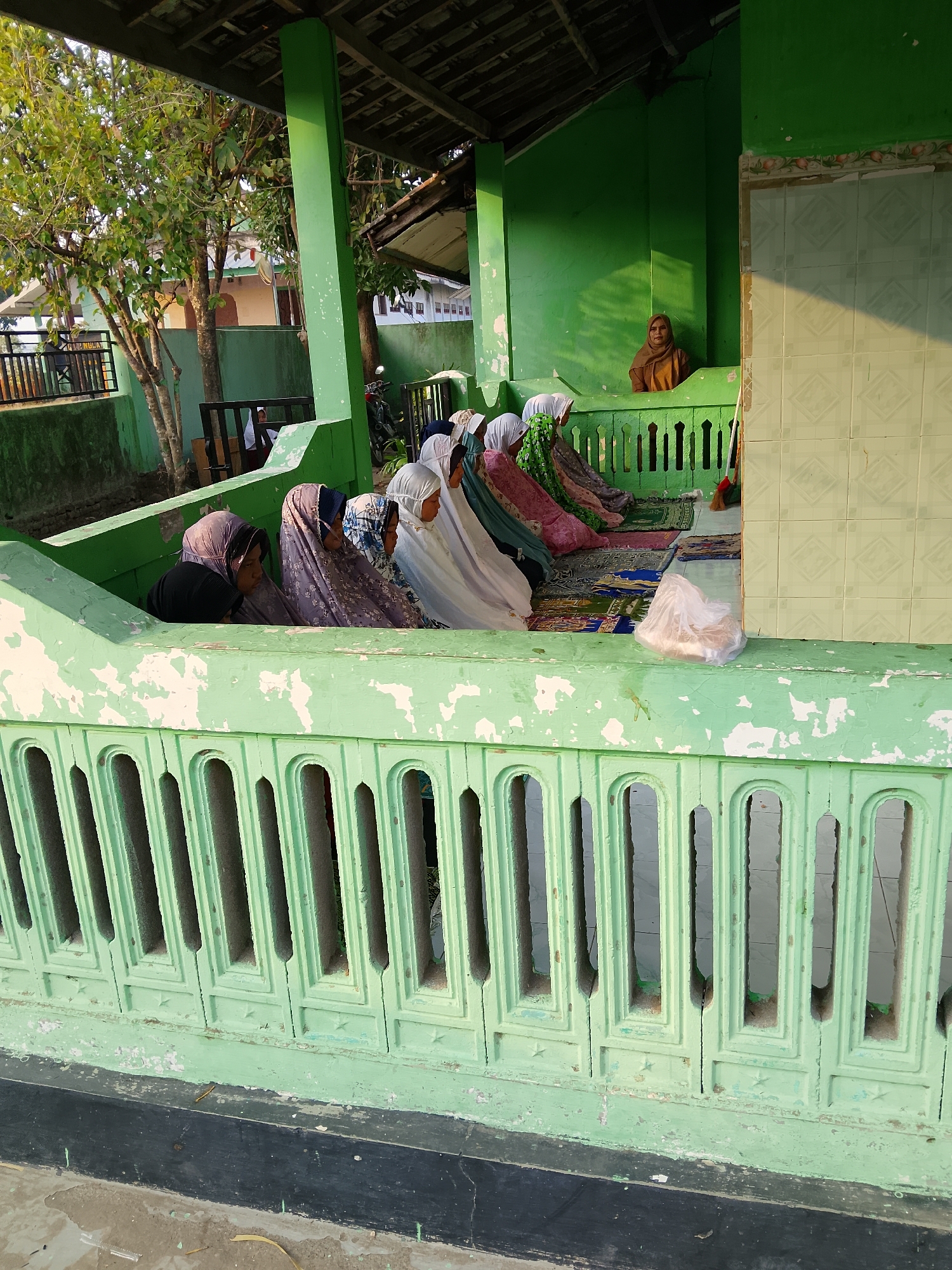 In the city of Kepuh, Indonesia, a group of women are engaged in prayer on prayer mats placed on the ground. They are seated in a covered area with green walls and a wooden roof structure. A single woman, wearing a brown headscarf, is visible sitting in the background, seemingly observing. The foreground features a weathered light green railing with a patterned design. The overall environment suggests a religious or spiritual gathering space, possibly part of a mosque or a similar sacred site. The lighting indicates it is either early morning or late afternoon.
