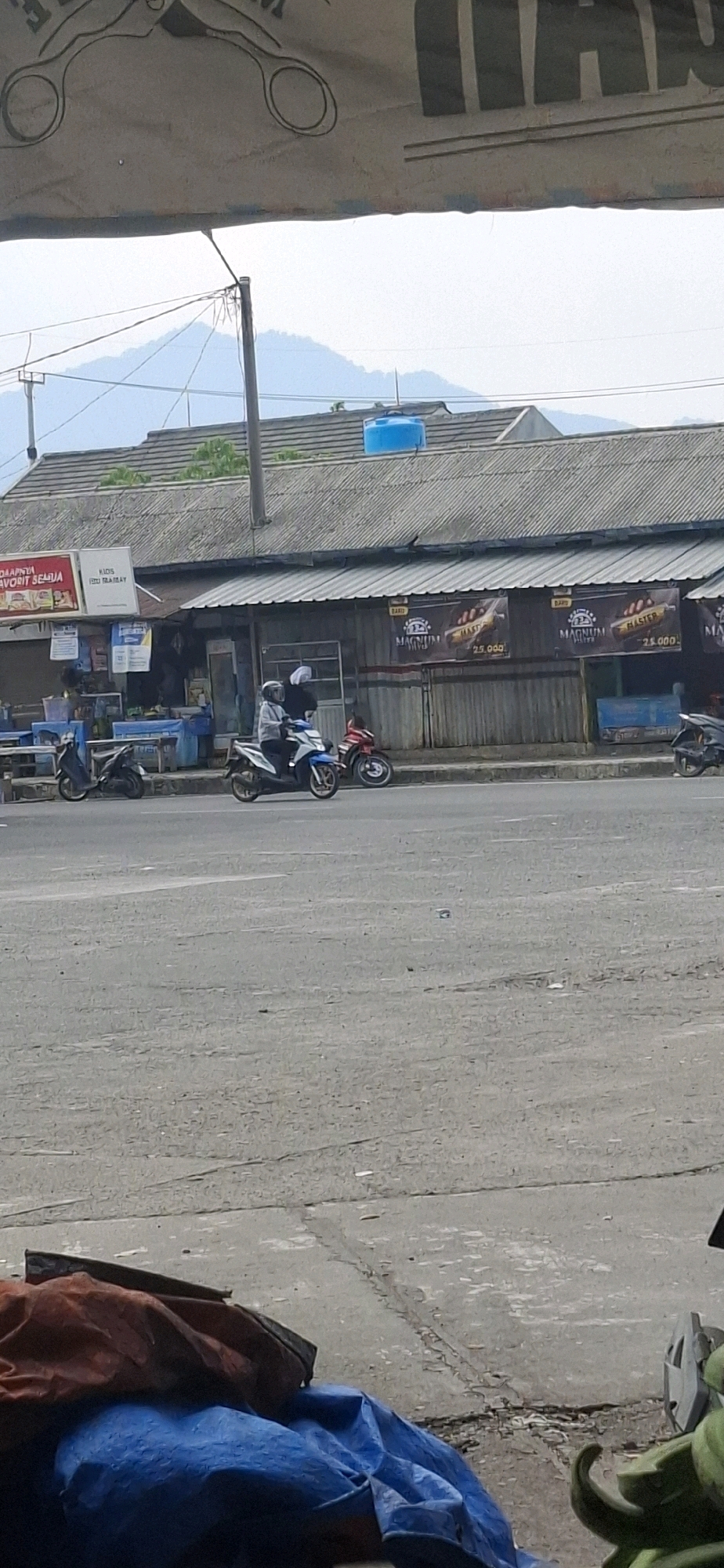 This is a vertical, medium-shot, outdoor photograph taken on a street in Sirnagalih, Indonesia. In the foreground, the edge of a street is visible with some debris. In the middle ground, there is a street with several motorcycles and two people. One person is riding a white and blue motorcycle, wearing a helmet and white clothing. Another person is standing behind the rider, wearing a white hijab. A red motorcycle is parked behind them, and another dark-colored motorcycle is parked to the left. Beyond the street is a building with a corrugated metal roof and advertisements on its facade. In the background, there is a mountain range under a hazy sky. The overall scene depicts a typical street view in a town or city.
