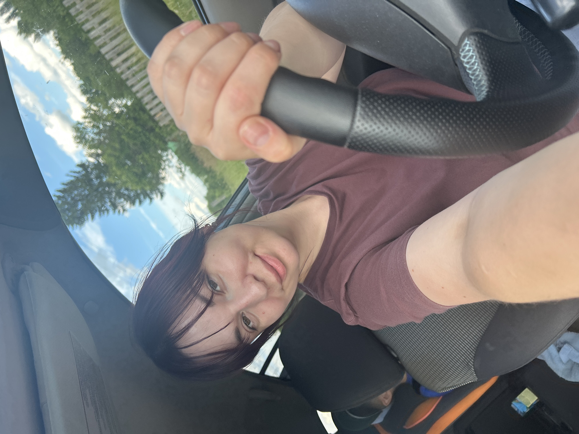 The image shows a woman sitting in the driver's seat of a car, holding the steering wheel. She has dark hair and is wearing a maroon t-shirt. Her face is turned towards the camera, and she is smiling. The background is out of focus, but appears to be a sunny day with green trees and a blue sky visible through the car's windshield. The interior of the car is dimly lit. There is no visible text in the image. The overall impression is of a casual, personal snapshot taken while driving.