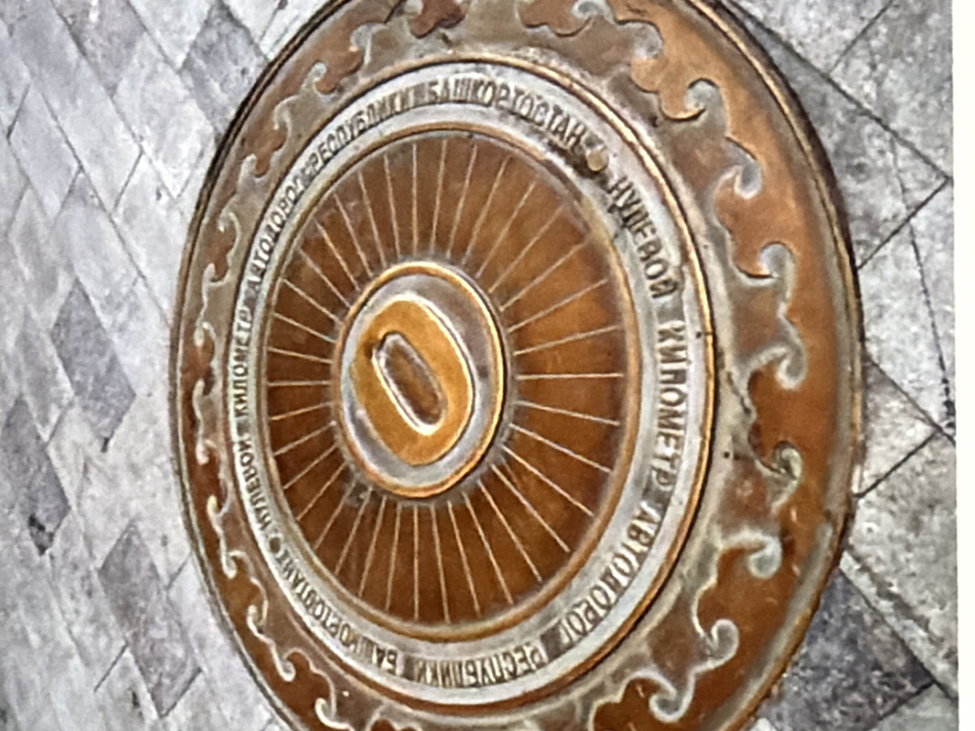 A close-up, overhead view captures a circular bronze plaque embedded in a cobblestone surface. The plaque is adorned with raised lettering that encircles a central design. The text, appearing to be in Russian, reads "КИЛОМЕТР АВТОДОРОГ РЕСПУБЛИКИ БАШКОРТОСТАН" which translates to "Kilometer of the Roads of the Republic of Bashkortostan." The central design features radiating lines emanating from a hollow oval shape in the very center, all rendered in bronze with a golden hue. The plaque is set into a ground composed of uniform, grey, rectangular cobblestones arranged in a diagonal pattern. The overall image presents a static, outdoor scene of a landmark.