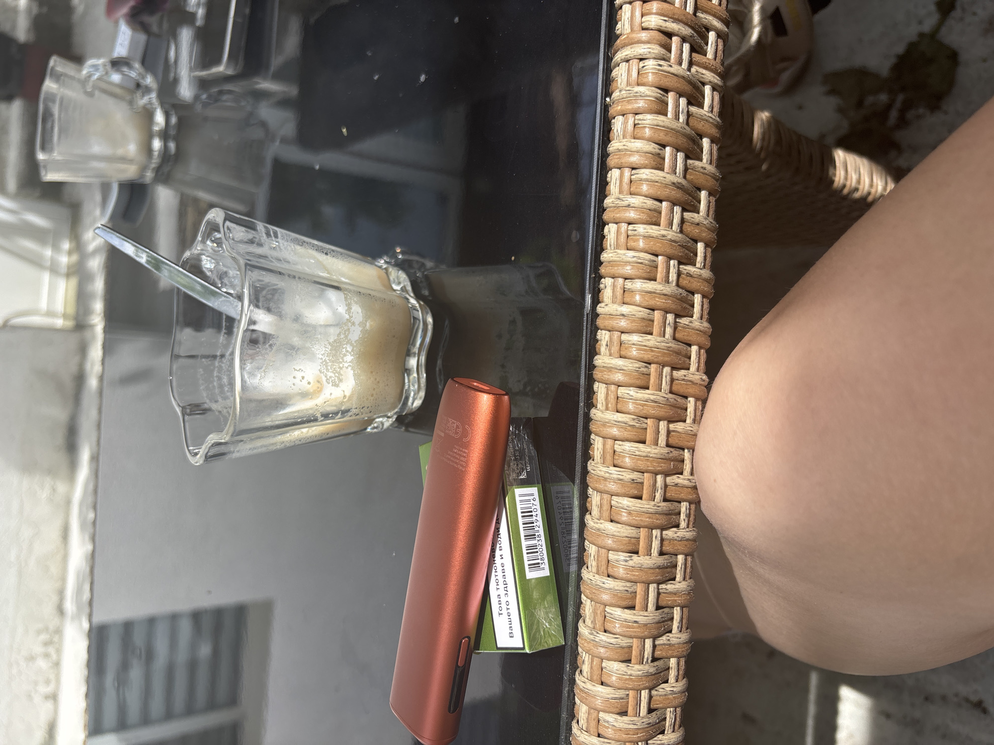On a glass-topped table with a woven edge sits a mostly empty glass coffee cup with a spoon inside, an orange electronic smoking device, and a pack of green cigarettes. A person's thigh is visible next to the table. The location is Unknown, Unknown.