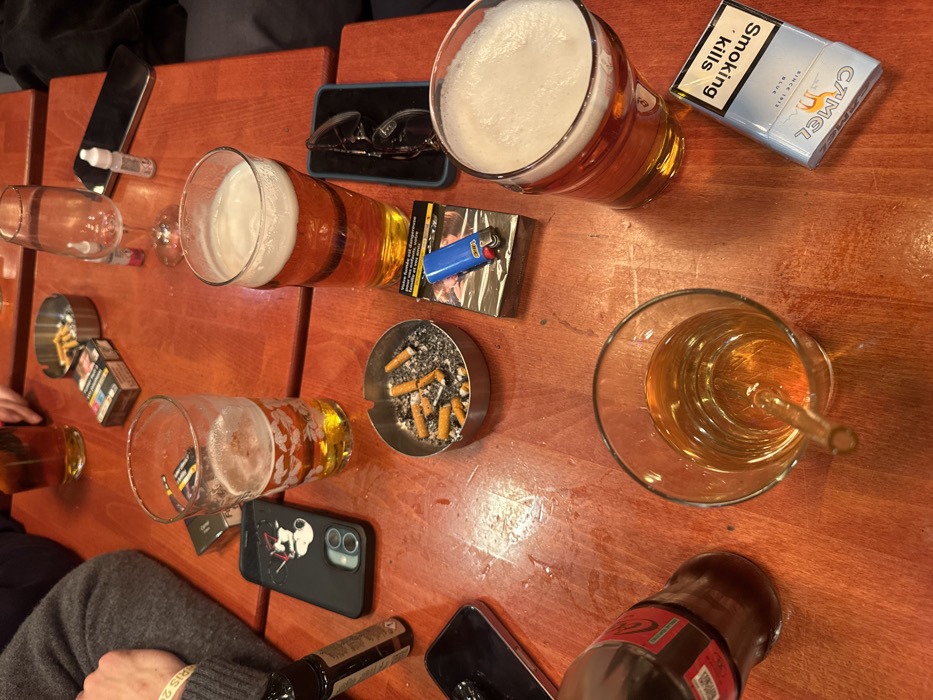 The image displays a reddish-brown wooden table in an indoor public establishment located in Vincennes, France. The scene is presented from an overhead perspective, focusing on various items associated with a social gathering involving multiple individuals.
**Objects and Subjects:**
* **Beverages:** Four glasses containing amber liquid are present. Three are pint glasses, approximately half to three-quarters full, each topped with a white foam head, indicative of beer. The fourth is a clear glass tumbler containing amber liquid with a clear glass straw. A partially consumed brown glass bottle with a red label, identifiable as "Coca-Cola zero sucre," is also on the table. An empty stemmed wine glass stands in the upper right section.
* **Smoking Paraphernalia:** Two small, circular metallic ashtrays are visible. One is filled with numerous cigarette butts and ash. The other, in the upper section, contains fewer butts. Three cigarette packs are present: one largely obscured, another partially wrapped in plastic with a blue Bic lighter resting on top, and an unopened "Camel Blue" pack oriented upside down, displaying the English health warning "Smoking Kills."
* **Electronic Devices:** A black smartphone encased in a black cover featuring a white graphic design and dual camera lenses is centrally located. Another black smartphone without a case is visible in the lower left. A black vape pen or e-cigarette device stands upright on the left side.
* **Personal Items:** A pair of black-framed eyeglasses is positioned in the middle right section. A small white plastic dropper bottle is near the empty wine glass.
* **Human Elements:** A portion of a person's left arm, clad in a grey long-sleeved garment, is visible in the upper left. A white wristband displaying "PARIS 2024" is on this arm. A dark fabric, likely part of another person's clothing, is partially visible in the upper right.
**Actions and Interactions:**
The collective presence of multiple drinks, full and used ashtrays, and various personal items indicates that several individuals are actively consuming beverages and tobacco products in a social setting. No explicit actions are depicted, but the arrangement implies ongoing interaction and leisure.