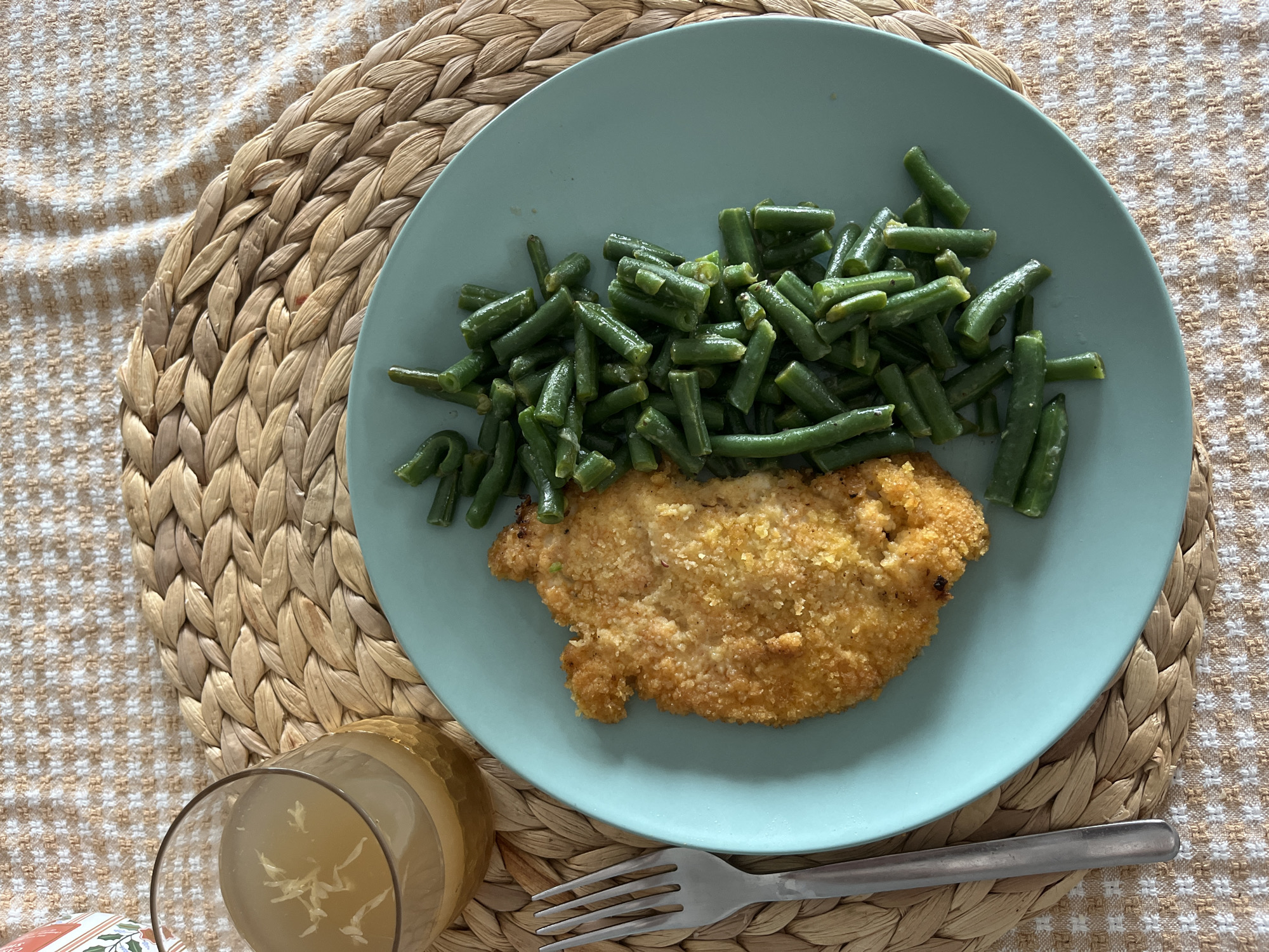 The image displays a meal set on a woven placemat atop a patterned tablecloth. A light teal-colored round plate occupies the center, holding a breaded, golden-brown cutlet on the left and a pile of cooked green beans on the right. To the upper left of the plate, a textured glass contains a pale yellow liquid with several small, white, shredded particles floating within it. A silver-colored metal fork is positioned vertically to the left of the plate. The placemat is made of light brown woven material, and the underlying tablecloth features a repeating beige and white checkered pattern. The scene depicts an indoor dining setting. The specific location in Ramat Gan, Israel, cannot be determined from the visual information provided.