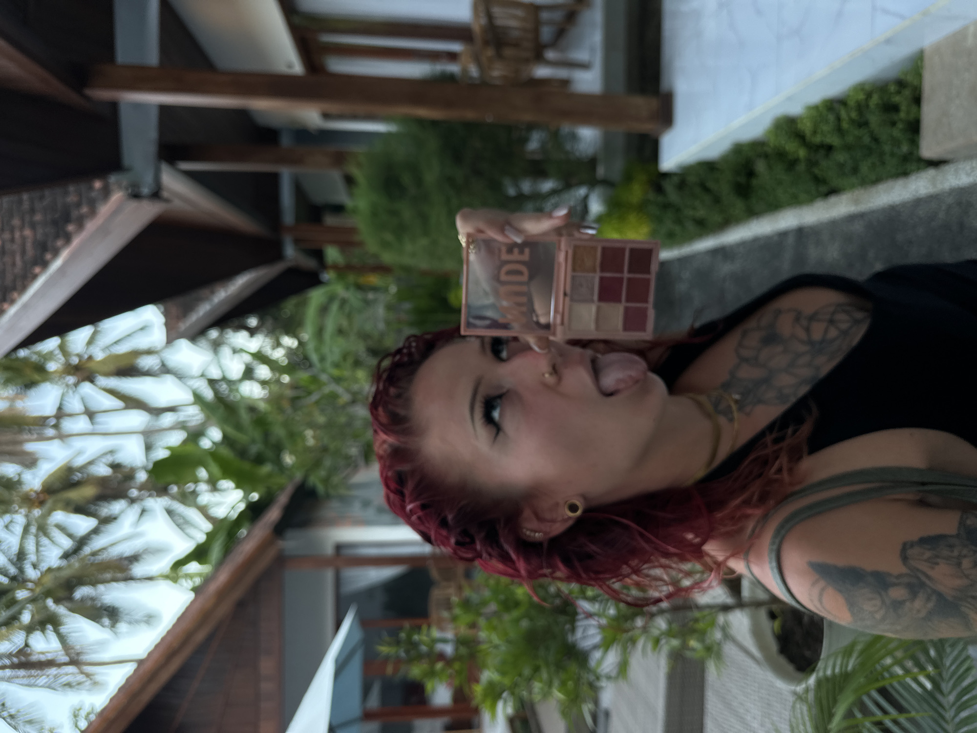 A woman with red hair and tattoos is holding a makeup palette in what appears to be Nusa Penida, Indonesia. She's sticking out her tongue. The background includes greenery and what looks like a building or resort structure, including the distinctive pitched roofs common in Balinese architecture.