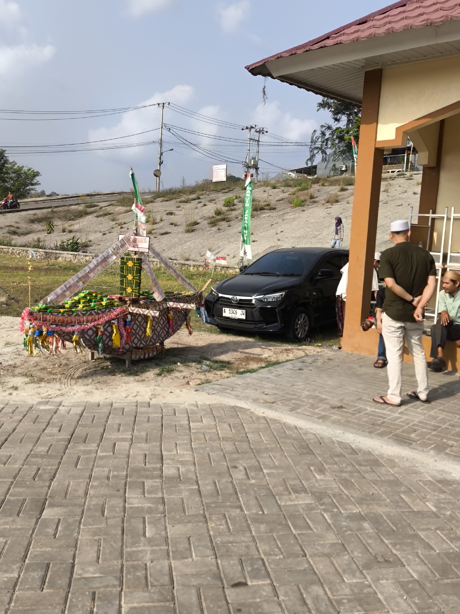 The image depicts an outdoor scene in Cilegon, Indonesia. In the foreground, a decorated structure resembling a boat, adorned with flags and what appear to be offerings, sits on sandy ground. A paved walkway with interlocking brick patterns runs through the foreground. To the right, a building with a brown and yellow facade and a reddish-brown tiled roof is visible. Several people are present. One individual, wearing a dark green shirt and white pants and a white cap, stands with his back to the camera. Another person, wearing a light green shirt and a straw hat, is seated. A black car is parked near the decorated structure. The background shows a sloped embankment with sparse vegetation, utility poles and wires, and a blue sky with scattered clouds. A person is seen riding a motorcycle in the distance on a road above the embankment.