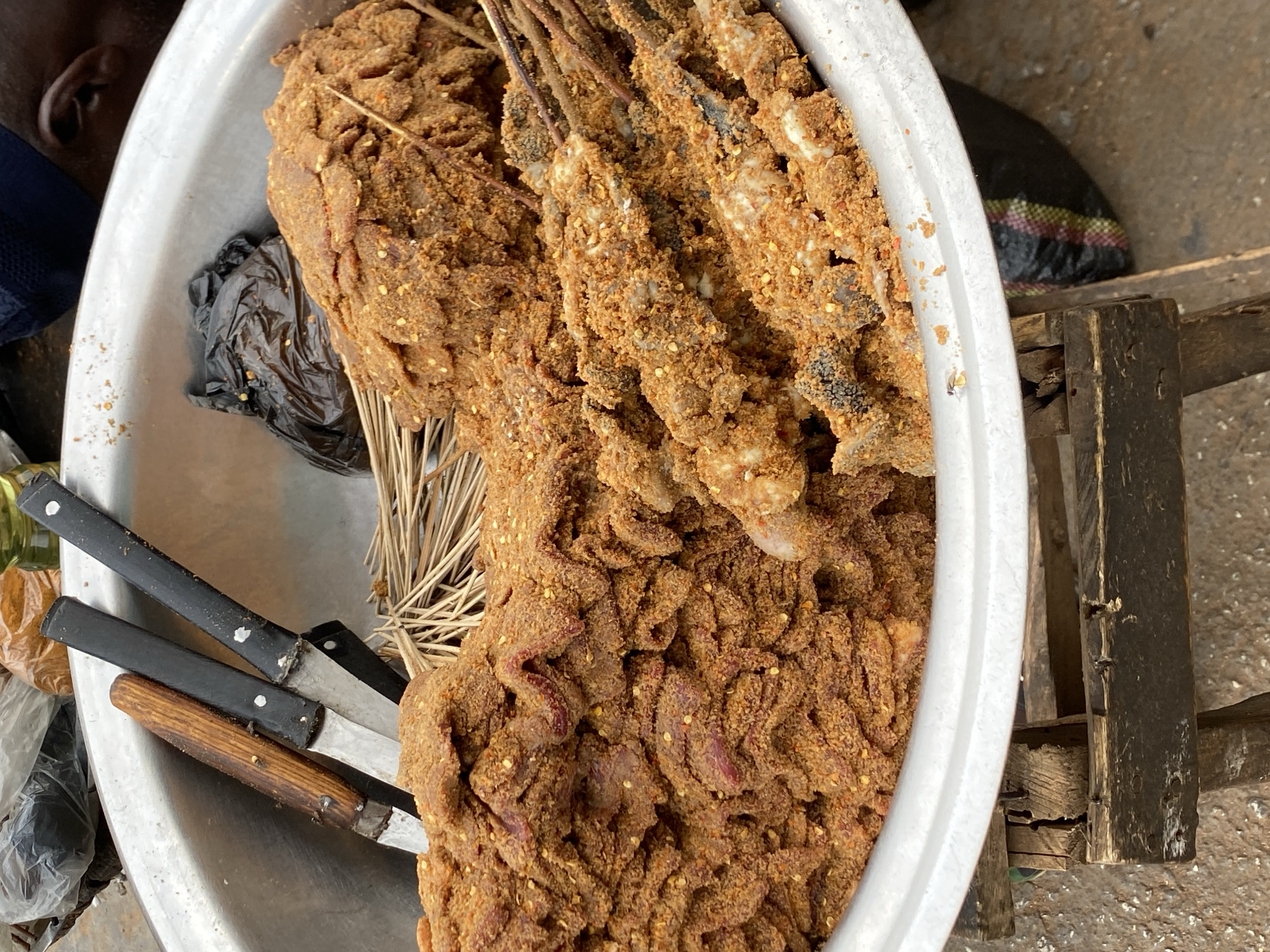 An overhead shot captures a large, silvery metal basin brimming with various spiced food items, likely at a market stall in Bukuru, Nigeria. The primary focus is a substantial quantity of raw or semi-prepared meat or fish, thoroughly coated in a coarse, reddish-brown spice mixture, possibly containing chili flakes and ground nuts.
Numerous pieces of food, resembling small fish or cuts of meat, are threaded onto thin wooden skewers and are also heavily encrusted with the spice blend, consistent with West African spiced meats like suya. To the left, a large, irregular mass of similarly spiced, thinly cut meat or offal is visible. A bundle of thin wooden sticks, likely for additional skewering or as fuel, lies beneath some of the food.
On the left side of the basin, three well-worn knives are laid out, two with black handles and one with a wooden handle. A crumpled black plastic bag is partially visible beneath the food. In the upper left corner, a partial view of a person's head, showing an ear and dark skin, is present. The metal basin rests on a rustic wooden stand, parts of which are visible on the right, along with a small black bag featuring a colorful stripe pattern. The scene depicts an active food preparation or vending setting. No discernible text is visible.