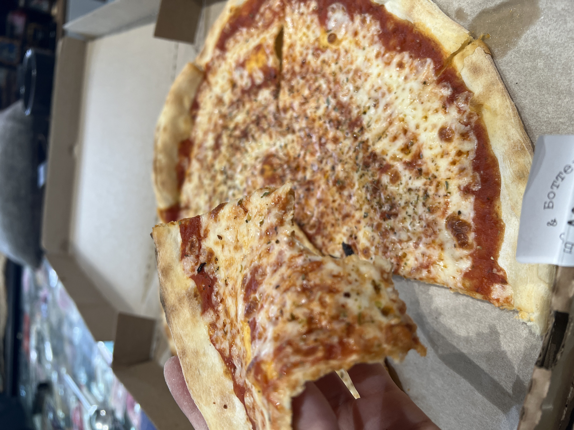 A partial pizza, mostly covered with melted white and yellowish cheese and a reddish tomato sauce, sits in an open cardboard pizza box. A slice of the pizza is being held up with a person's fingers. The box is sitting on a surface. In the blurry background to the left is what seems to be a shop or store with reflective surfaces. A paper with writing on it is to the right of the pizza. There is no indication of the location being Khimki, Russia, in the image.