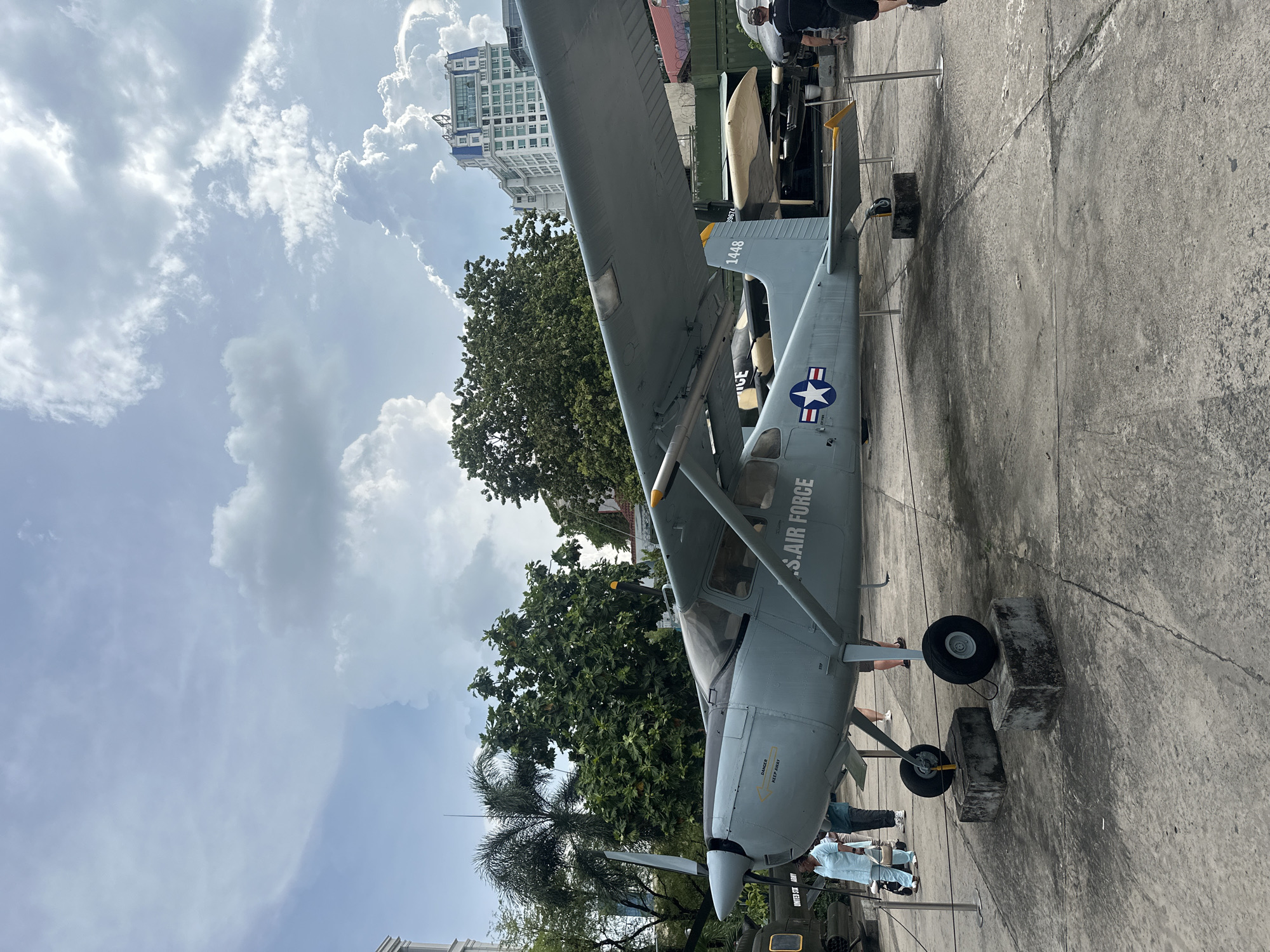 A grey U.S. Air Force aircraft, identified by "U.S. AIR FORCE" lettering and a U.S. star insignia, is on display outdoors in what appears to be a museum setting in Thủ Đức, Vietnam. The plane is positioned on concrete blocks, likely for preservation or display. Trees are visible in the background. The sky is overcast with buildings also in the background. People appear to be observing the aircraft, although their activities are not clear due to the image perspective.