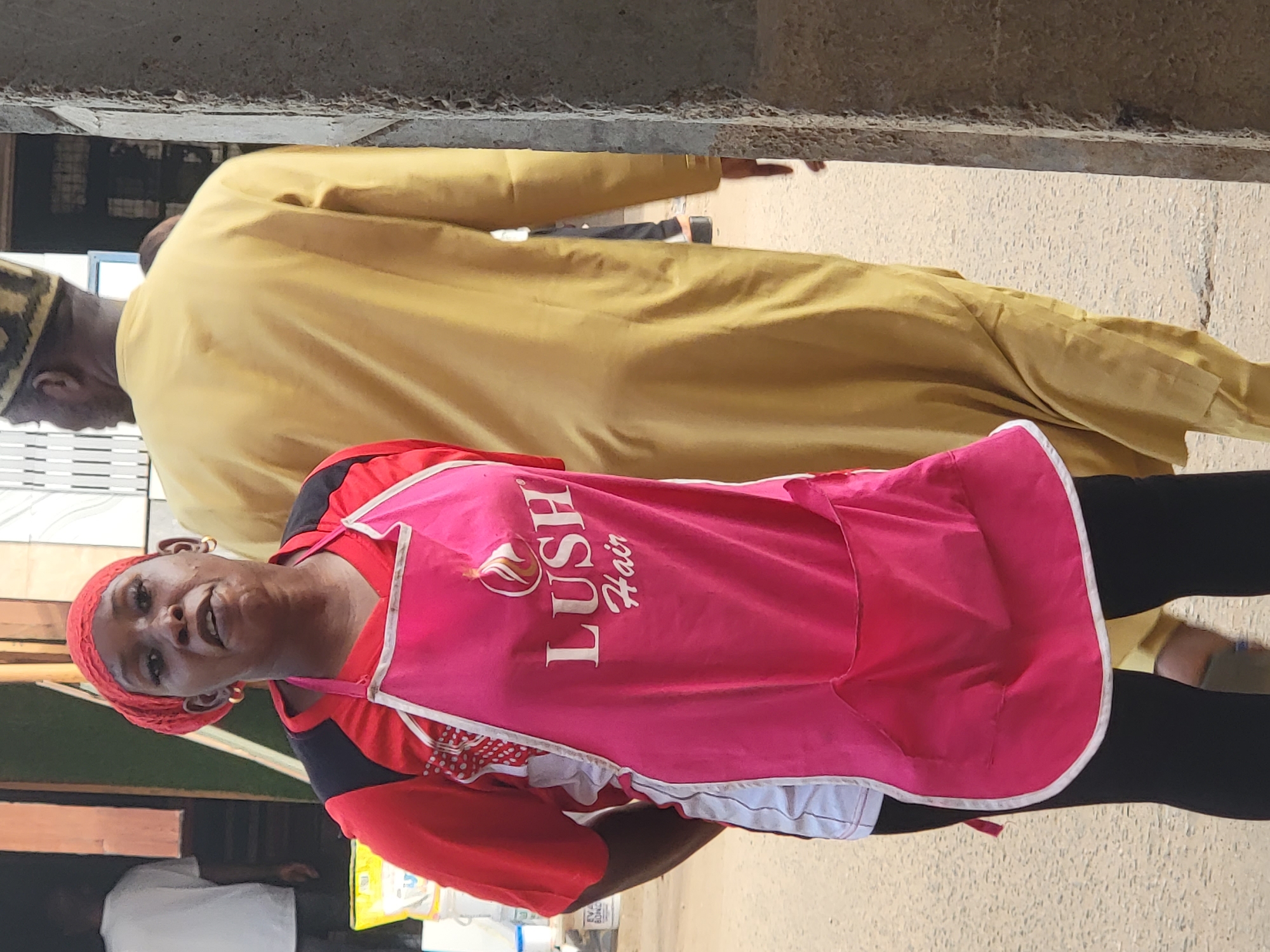 A woman wearing a pink apron with "LUSH Hair" printed on it stands in Maiburiji, Nigeria. She is wearing a red headscarf and a red shirt with black and white accents. A man in a yellow robe and a patterned hat is walking behind her. The scene appears to be in a busy commercial area.
