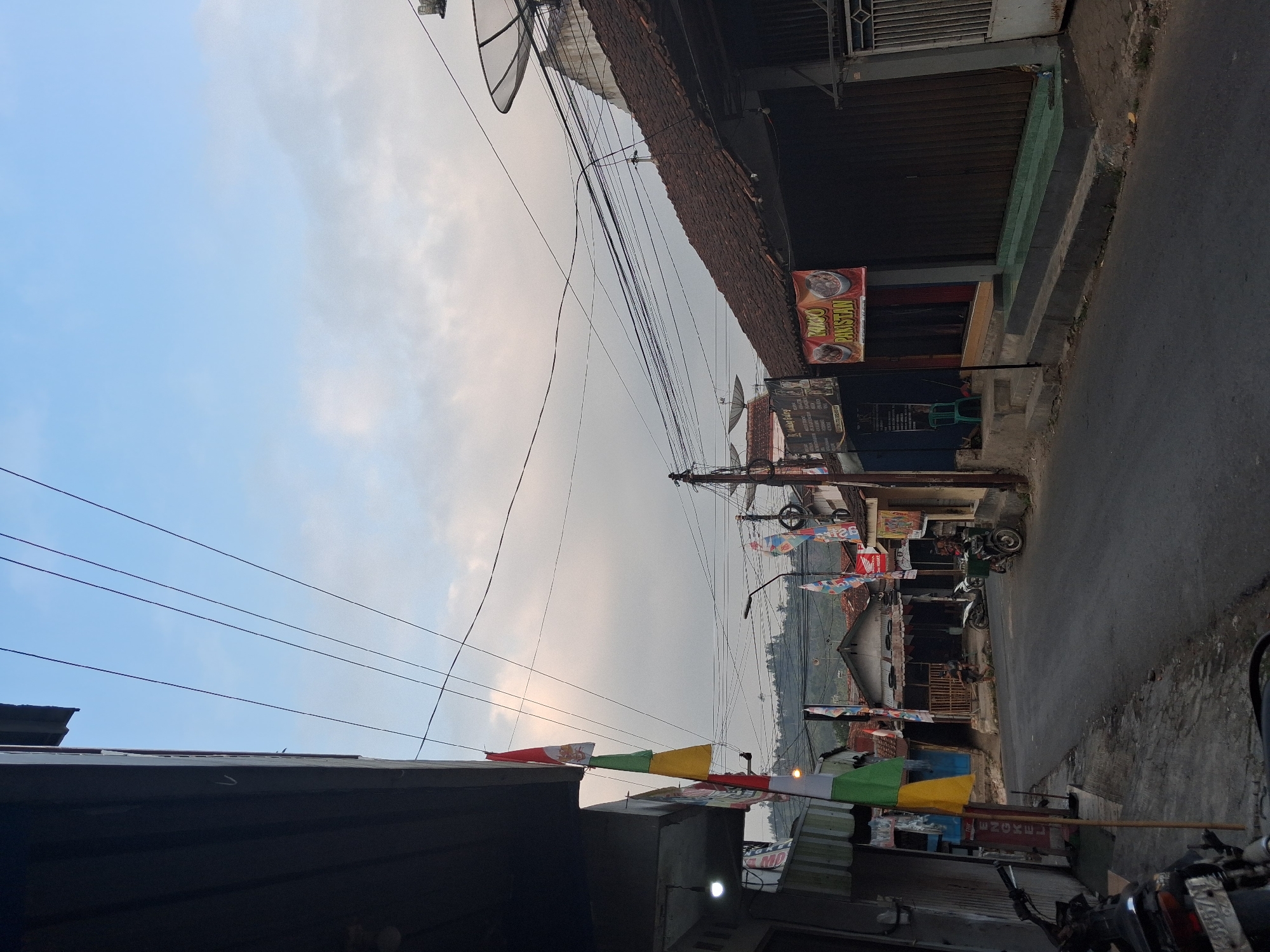 The image depicts a street scene in Bantarkawung, Indonesia, during daylight. A paved road runs through the center of the frame, lined by buildings and shops on both sides. Several motorcycles are parked along the roadside. Overhead, a complex network of electrical wires crisscrosses the sky, supported by utility poles. Decorative flags, predominantly red and white and green and yellow, are hung from poles and buildings. On the right side of the street, a building with a tiled roof features a banner advertising "BAKSO PAKISTAN." The sky above is partly cloudy, with a mix of blue sky and diffused light from the clouds. In the background, hills are visible. The scene appears to be a typical rural or semi-urban street in Indonesia.