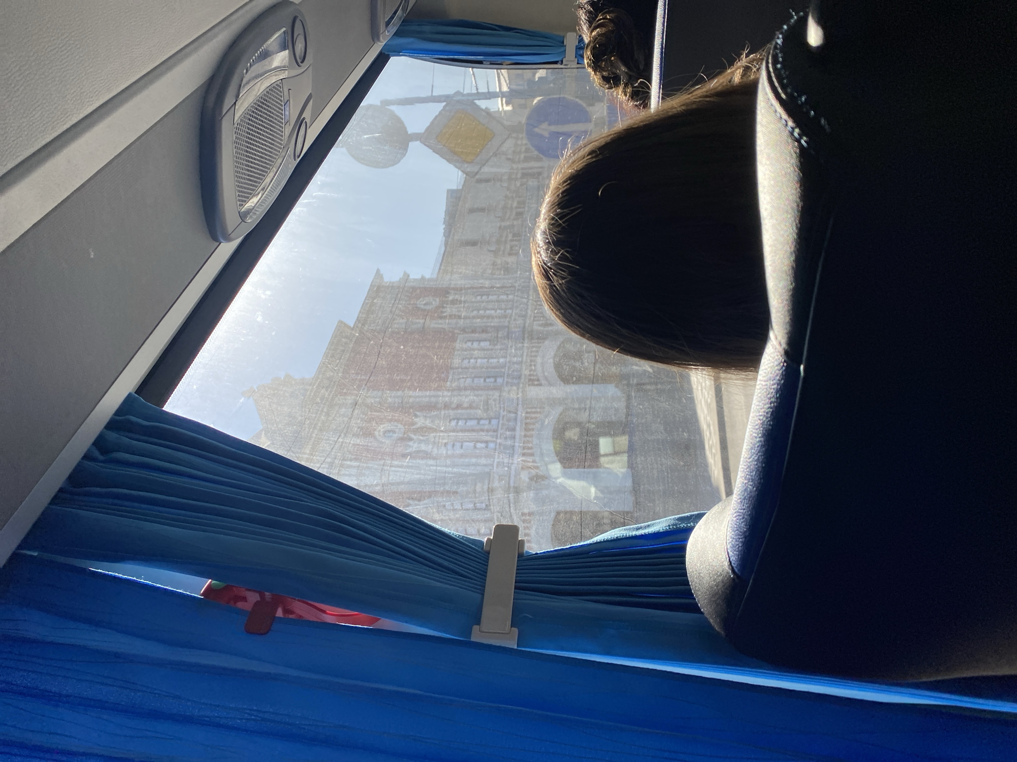 A view from inside a bus shows a street scene through the window. The window has blue curtains. Part of the back of a person's head is visible in front of the window. The reflection of a building is visible in the window. The street signs are also visible in the window. The building in the reflection has brickwork and is multi-story. Based on the architecture of the building, the street signs, the bus seat, and overall atmosphere, the location of the image is Moscow, Russia.