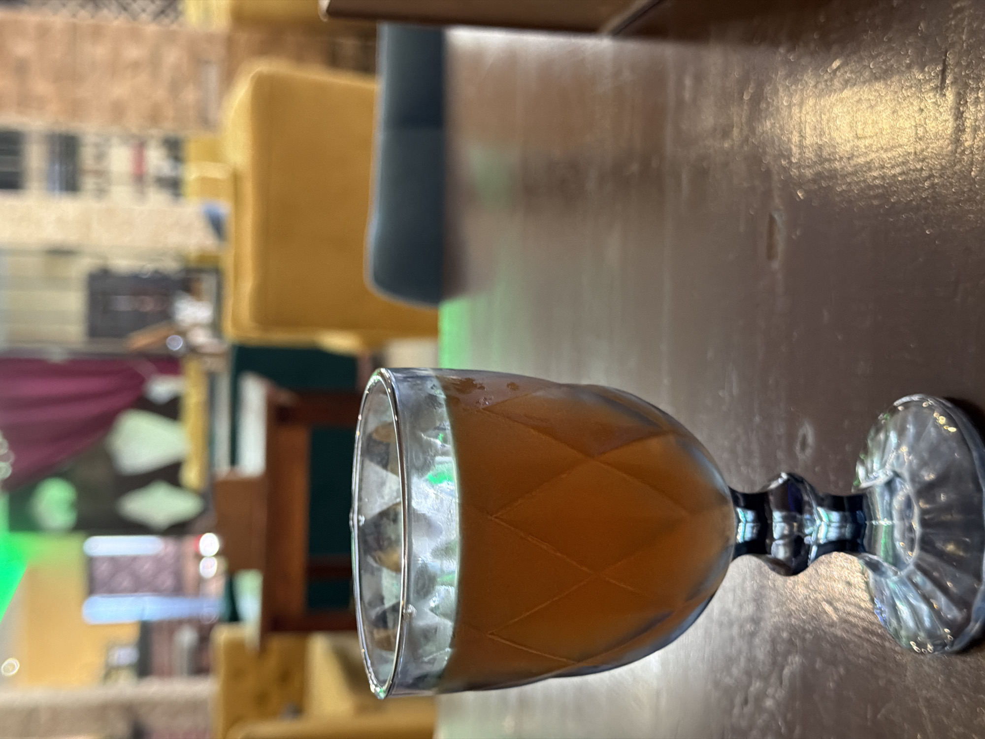 A stemmed glass filled with an amber-colored liquid sits on a dark wooden table in what appears to be a restaurant or cafe. The glass has a faceted design. In the background, out of focus, are yellow chairs and tables, with a glimpse of a purple curtain and green lighting. The overall scene suggests an indoor setting, likely a public establishment. The location is the city of Хохряки, Russia.