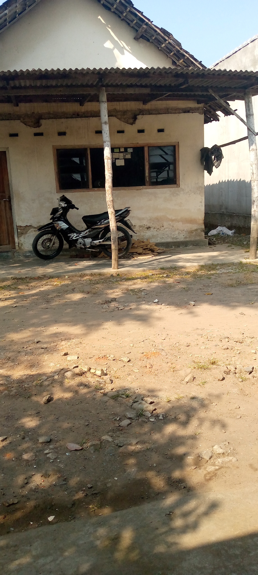 A black motorcycle is parked in front of a weathered, light-colored building in Sumberwono, Indonesia. The motorcycle, with visible "SUZUKI" branding, is positioned on a dirt and gravel surface, partially covered in shadows cast by overhead foliage. The building appears to have a corrugated metal roof supported by wooden posts. Two windows with dark frames are visible on the building's facade. To the right of the motorcycle, a pile of cardboard is stacked near the wall. Above the windows, the wall has small, dark rectangular vents. On the right side of the building, dark clothing hangs from a line. The ground in front of the building is a mix of dirt, small stones, and patches of sparse grass. A white plastic bag lies on the ground further to the right. The sky above is clear and blue.