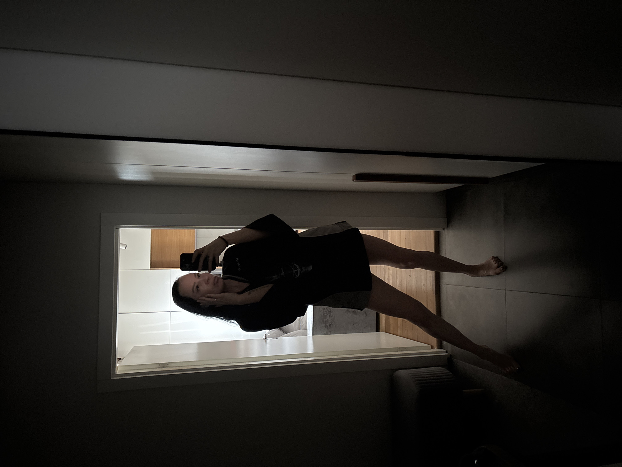 A woman is standing in a doorway taking a selfie. The doorway leads to a brightly lit room with white cabinets and light-colored walls. The floor outside the doorway is dark tile. The woman is wearing a dark shirt and light shorts, and she is holding a black phone in her right hand. Her left hand is near her face. The location is not explicitly determined by the image. It is impossible to confirm whether the location is Chaikovsky, Russia, based solely on visual information.