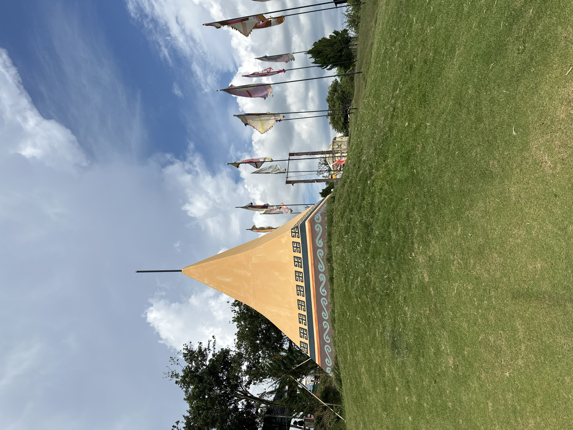 The image depicts an outdoor scene on a grassy hill, likely in the city of Thôn 5, Vietnam, given the context. The central focus is a large, orange, pyramid-shaped structure with decorative bands around its base, featuring geometric patterns and wavy designs. A thin, black pole extends vertically from the apex of the structure. To the right of the pyramid, a line of tall flagpoles displays various colorful flags, many appearing tattered or wind-worn. In the background, trees are visible, and a swing set with a wooden frame is present. The sky above is partly cloudy with patches of blue sky. The ground is covered in short, green grass. There are no visible people directly interacting with the central structure or flags in the foreground.