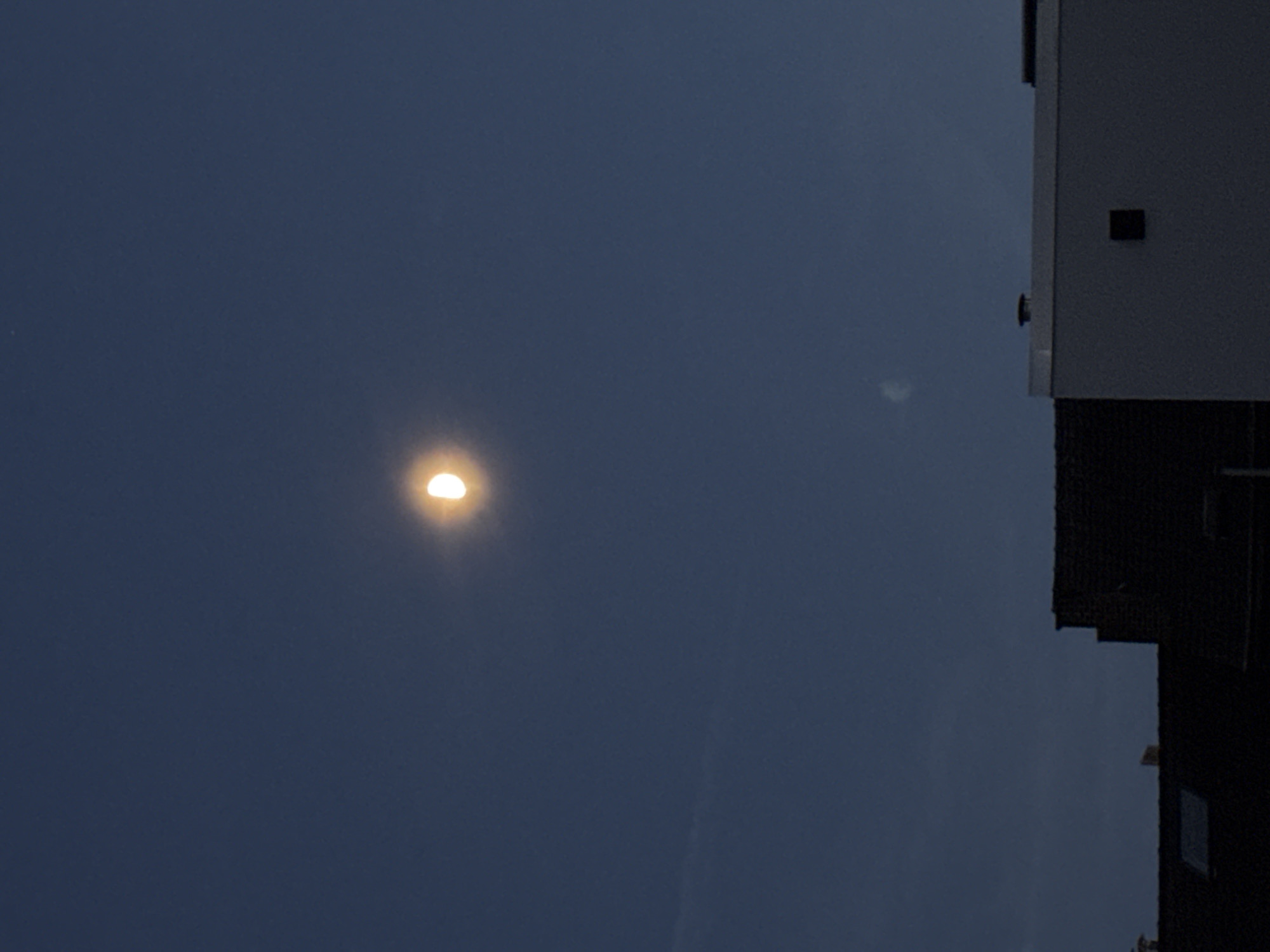 The image shows a sliver of the moon in a dark blue sky, above several buildings. The moon is illuminated on its right side, with the left side in shadow. One building in the foreground is dark and appears to be a residential building with a skylight. Another building to the right is white with a dark square window. The sky is clear with no clouds visible. The location is Molenbeek-Saint-Jean - Sint-Jans-Molenbeek, Belgium.