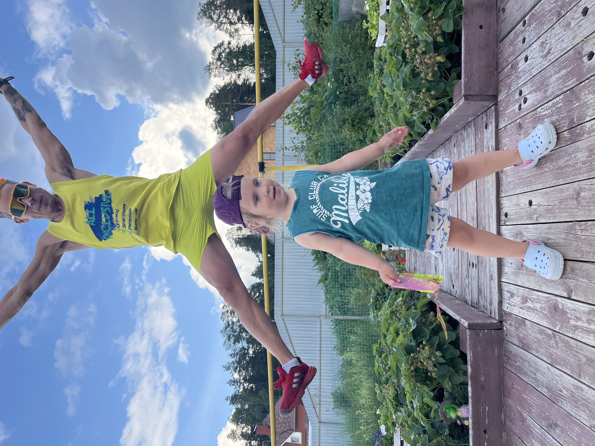 In a backyard setting, likely in the city of Chaikovsky, Russia, a man in mid-air performing a jumping jack is positioned directly above a young child. The man, wearing a yellow tank top and shorts, sunglasses, and red athletic shoes, is captured at the peak of his jump. The child, standing on a wooden deck, wears a green sleeveless shirt, patterned shorts, a purple baseball cap, and blue Crocs. The child holds a pink and yellow toy. Behind them, a garden bed with green plants is visible, and a wooden fence encloses the yard. The sky is blue with scattered white clouds. The overall scene appears to be a candid, playful moment outdoors.