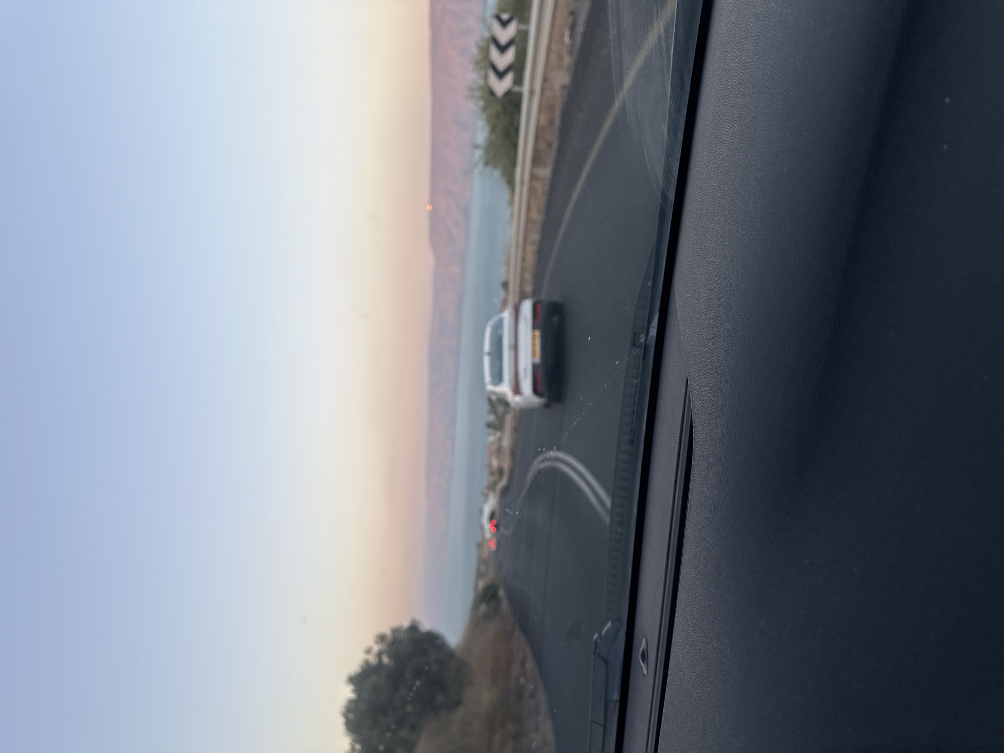 The image, taken from inside a vehicle in the city of Emek HaYarden Regional Council, Israel, captures a scenic view of a winding road leading towards a large body of water, likely the Sea of Galilee, at either dawn or dusk. The sky displays a gradient of soft blues and pinks, suggesting the golden hour.
In the foreground, the dark, textured dashboard of the car is visible, along with a portion of the windshield. A white sedan is driving ahead on the road, slightly to the right of the frame's center. Further down the road, two sets of red taillights indicate other vehicles. The road is asphalt with white lane markings, and guardrails with chevron signs are visible along the right side. To the left of the road, a rocky embankment covered with sparse vegetation rises, with a dark, leafy tree on its upper portion.
In the background, the expansive water stretches across the horizon, bordered by distant hills or mountains rendered in a muted pinkish hue. A small, bright orange dot, possibly the sun or a reflection, is visible above the distant hills. The overall impression is one of a serene, late-day drive through a picturesque landscape.