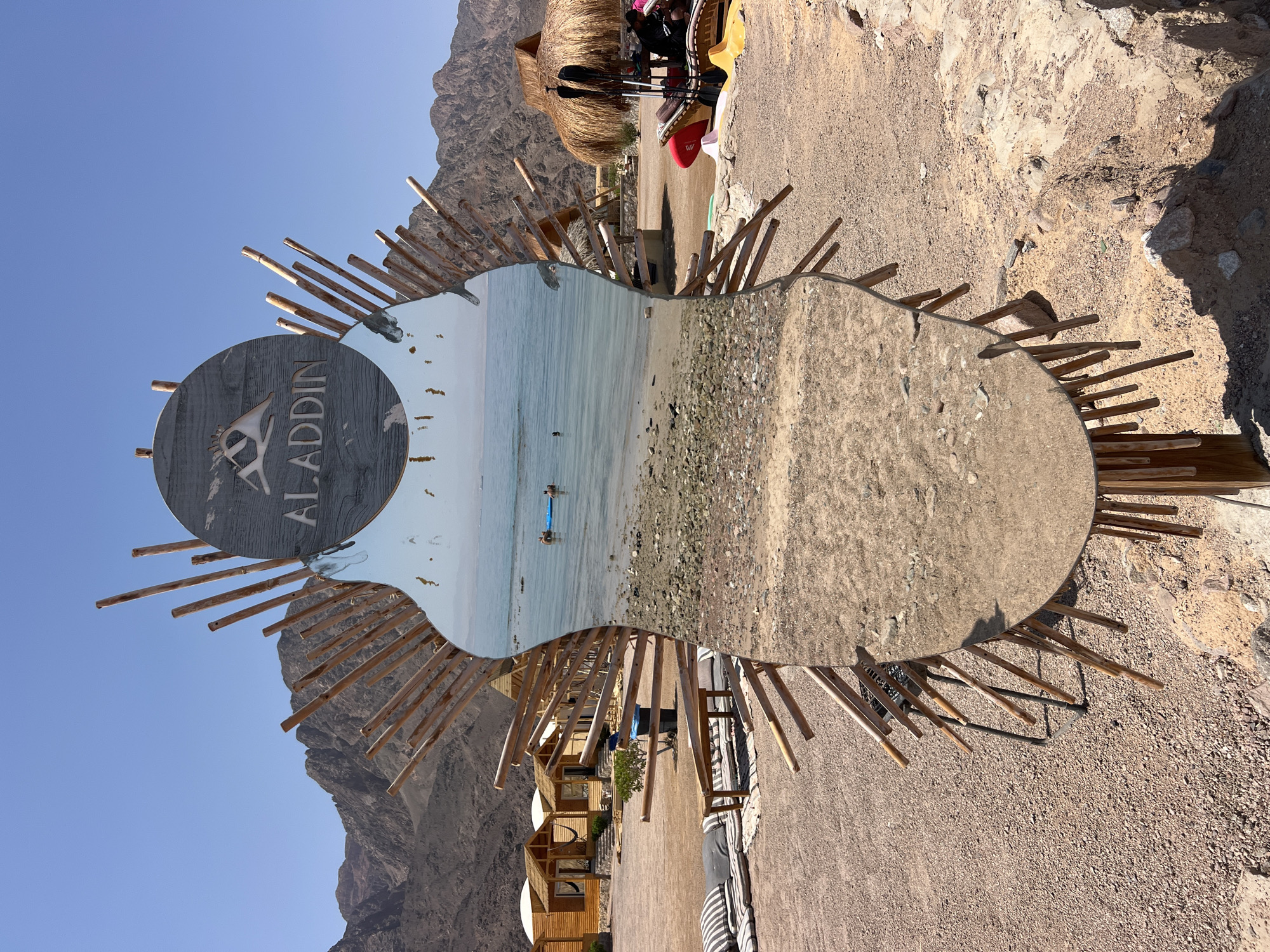 The image depicts a mirrored sign in the shape of a stylized human torso, adorned with bamboo poles, located at the Aladdin Beach Camp in Nuweiba, Egypt. The mirrored surface reflects a scenic view of the Red Sea, with calm, clear blue water, a sandy beach dotted with pebbles, and distant mountains under a bright, cloudless sky. Two individuals are visible in the water, one on a blue inflatable raft and another swimming. In the foreground, the beach camp is characterized by several wooden huts with thatched roofs, lounge areas with cushions and benches, and some recreational equipment like paddleboards and kayaks. The ground is covered in gravel and sand, typical of a desert coastal environment. The sign itself features the "ALADDIN" logo with a stylized camel silhouette.