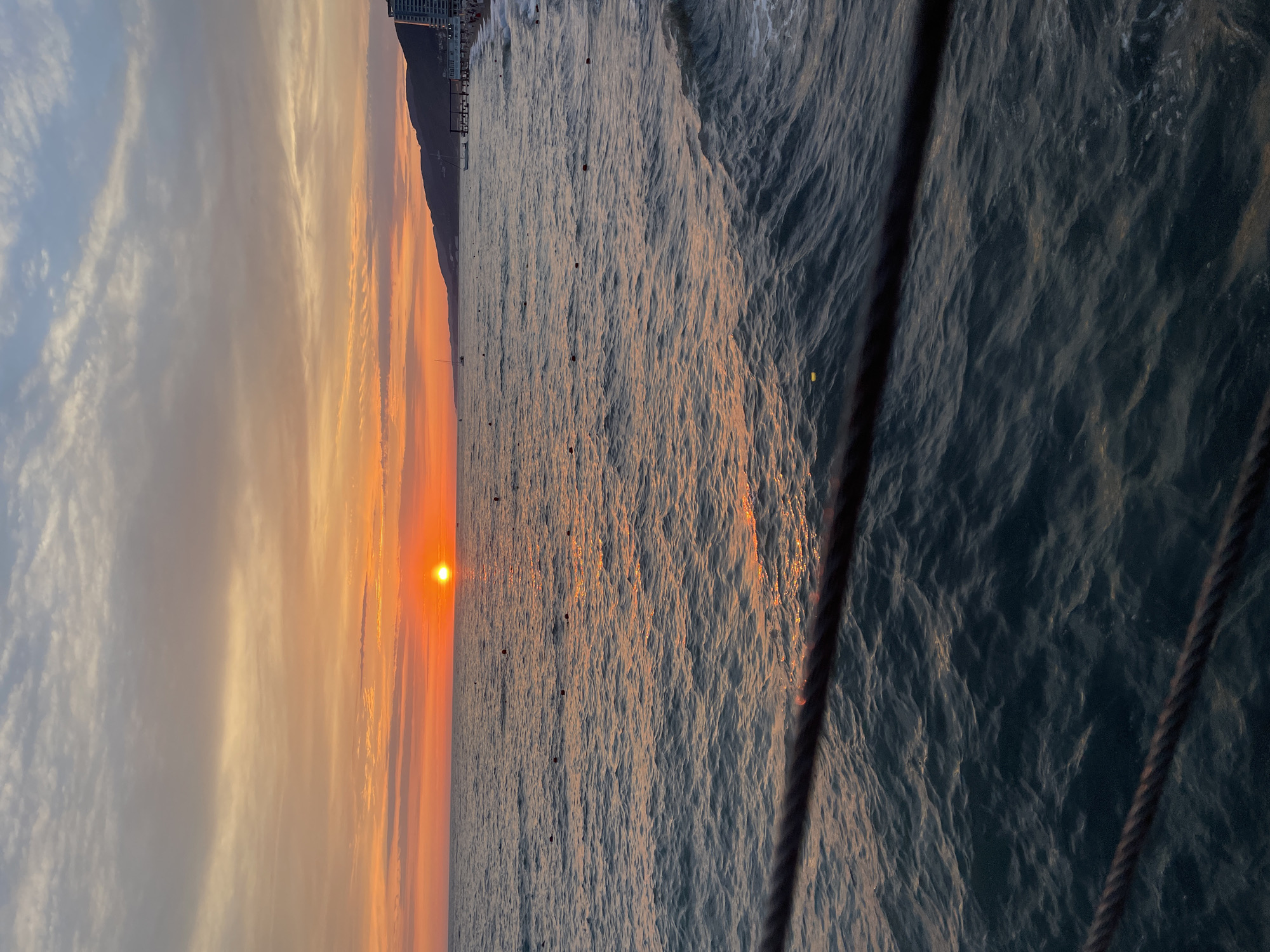 The image captures a scenic sunset over a vast body of water, likely the ocean, with a coastline visible in the distance. The sun, a bright orange orb, is low on the horizon, casting a warm glow and shimmering reflections across the rippling surface of the water. The sky is a gradient of colors, transitioning from pale blue at the top to soft orange and pink hues near the horizon, with wispy clouds scattered across the expanse.
The coastline features a mountain or cliff face that slopes down towards the sea, and buildings, possibly hotels or apartments, are perched along its edge, suggesting a coastal town or city. The water itself is a deep blue-gray, with visible waves and whitecaps, indicating a dynamic sea. Several dark, small objects are scattered across the water's surface, which could be buoys or small boats. In the foreground, a thick rope or cable runs diagonally across the frame, partially obscuring the water and adding a sense of perspective, as if the photo was taken from a pier or a ship. The overall mood of the image is tranquil and serene, evoking the beauty of a coastal sunset. The time of day is clearly sunset, and the weather appears to be clear with some cloud cover. There is no visible text in the image.