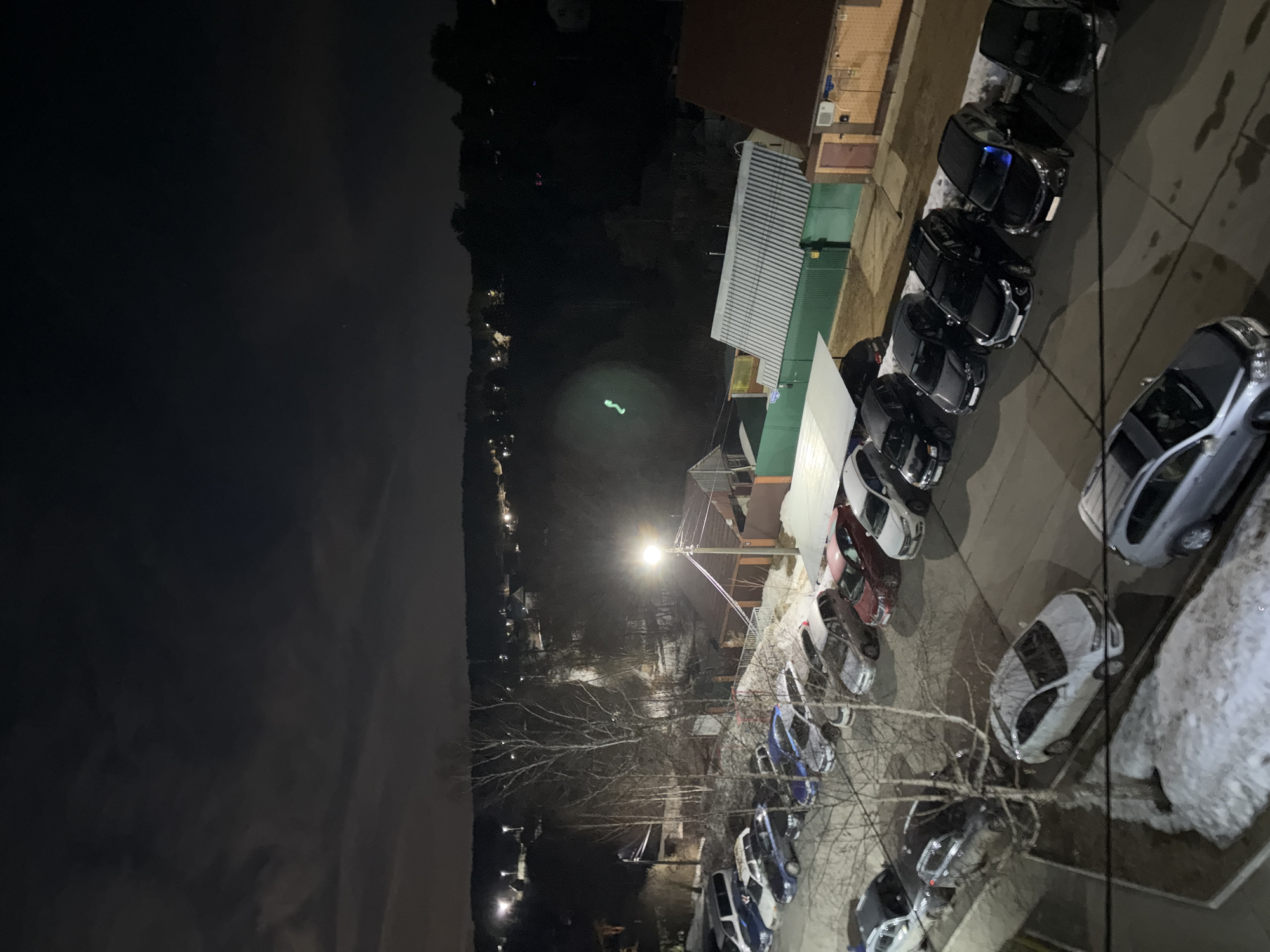 The image shows an elevated nighttime view of Chaikovsky, Russia, featuring a line of parked cars along a street and a densely vegetated area interspersed with streetlights and buildings in the background. The sky is dark and cloudy. Snow is present on some of the parked cars and in patches along the street, indicating a cold climate. A street light illuminates the parking area and the surrounding area, with bare tree branches visible between the cars and the light source.