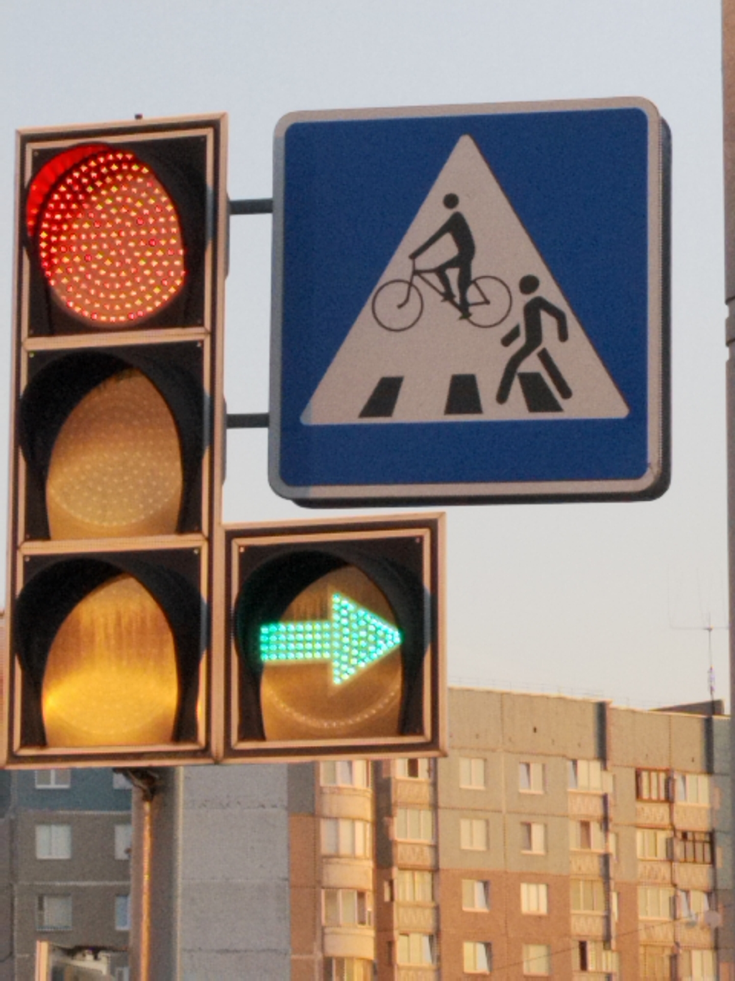 The image shows a traffic light with a red, yellow, and green light, and a pedestrian crossing sign in Hrodna, Belarus. The traffic light is red, indicating that vehicles must stop. The pedestrian crossing sign shows a silhouette of a person walking and a cyclist, indicating a shared pedestrian and cyclist crossing. In the background, there is a residential building. The sky is clear and the weather appears to be mild.
