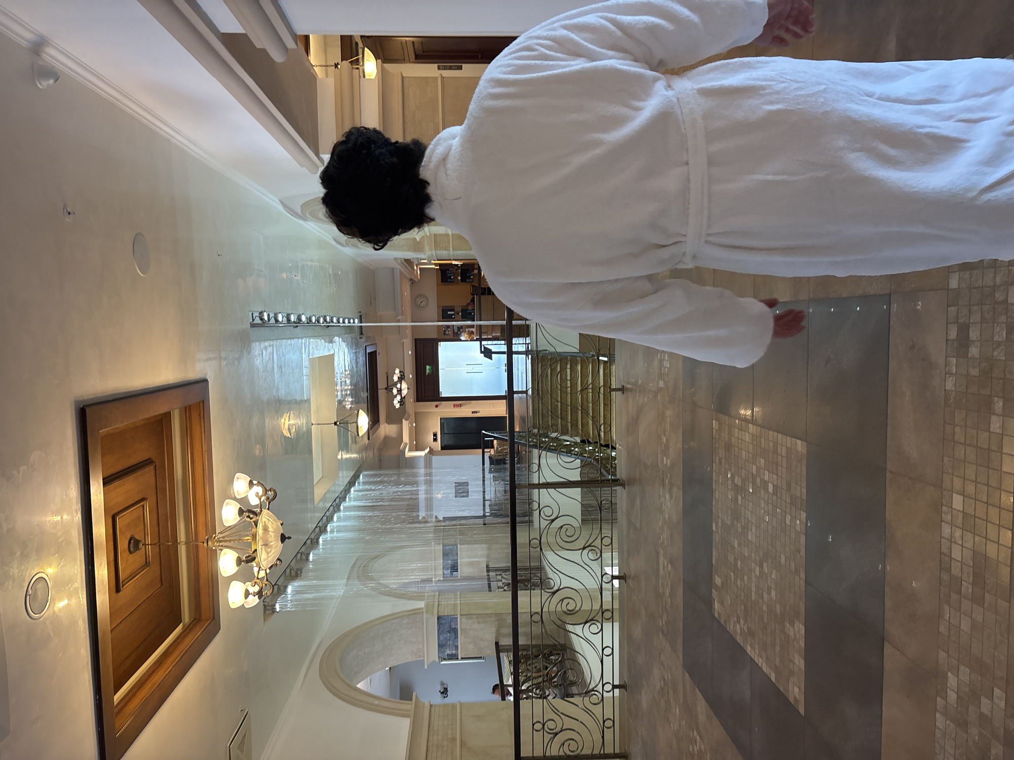 The image shows the interior of a building, possibly a hotel or spa, with a person wearing a white bathrobe walking away from the camera down a tiled hallway. In the background, there is a water feature cascading down from the ceiling. The architecture includes arches and ornate metal railings. There are chandeliers and pendant lights, and a wooden ceiling inset with a lighter wooden square. The floor is tiled in a pattern of large and small tiles. The scene is well-lit, suggesting an indoor environment. The location in the city of Razliv, Bulgaria is not visually evident from the provided image.