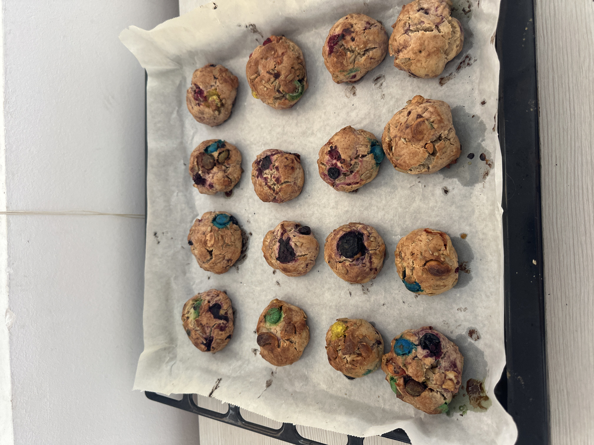 A batch of freshly baked cookies sits on a baking sheet lined with parchment paper. The cookies, appearing to be a rustic, homemade variety, are a golden brown color and some have visible chunks of colorful candies, possibly M&Ms, and other inclusions like seeds or dried fruit. They are arranged in a grid-like pattern across the baking sheet, with 16 cookies visible in total. The baking sheet itself is dark-colored and sits on a light-colored countertop. The background shows a white tiled wall. There are no people or animals present in the image. The scene appears to be set in a kitchen environment. The image does not contain any visual information that can be used to determine the location in the city of Budva, Montenegro.