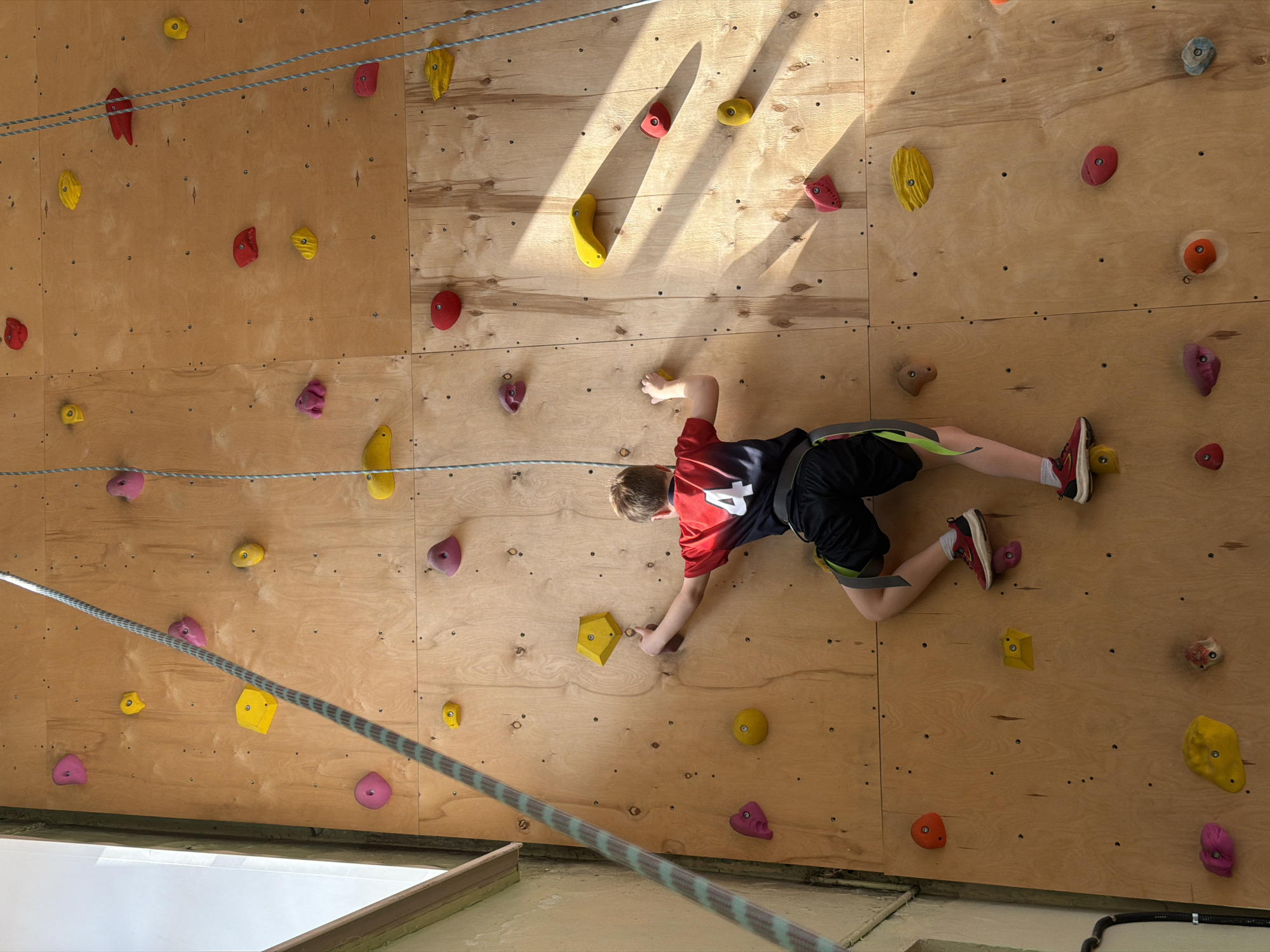 A young person is rock climbing on an indoor wall. The climbing wall is made of plywood and has various colored holds attached. The climber wears a red and black shirt, dark shorts with green trim, and red shoes. The climber's limbs are extended as they grip and step on the holds. Two ropes hang in front of the wall. The background shows signs of indoor construction. There is no location information (Izhevsk, Russia) visible in the image.