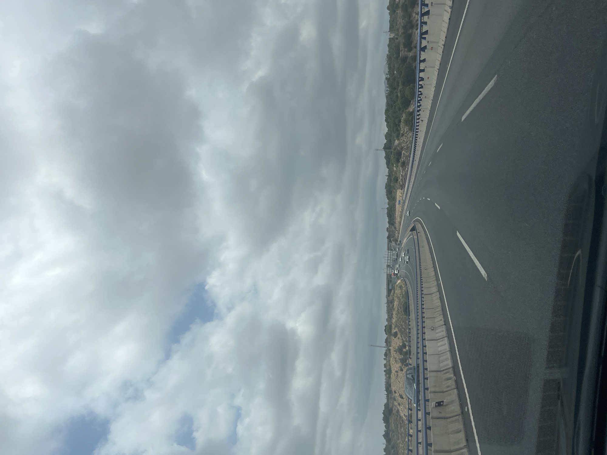 The image shows a view from a moving vehicle on a multi-lane highway. The highway curves to the right in the distance. The sky is heavily overcast with thick, gray clouds. The landscape on either side of the highway consists of dry, arid terrain with sparse vegetation and some hills. Concrete barriers and metal railings line the sides of the road. Several cars are visible on the highway, driving in the same direction as the viewpoint. The overall scene suggests a drive through a somewhat desolate, possibly rural or suburban, area under a cloudy sky. The provided information suggests the location is the city of Elx / Elche, Spain.