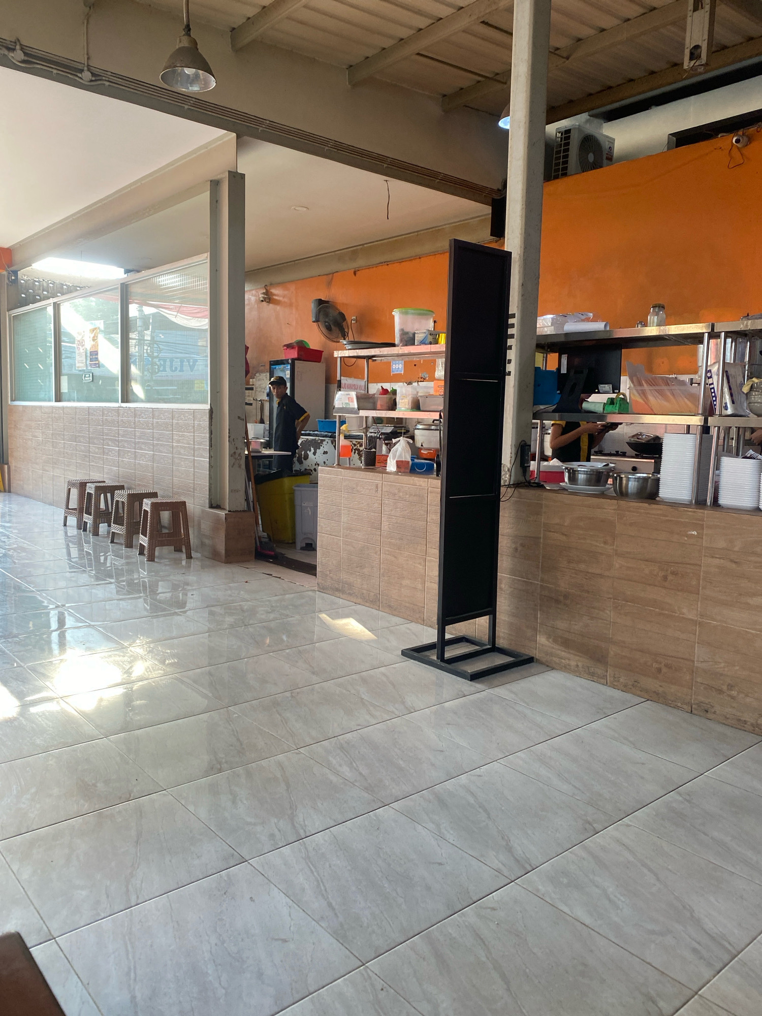 The image depicts the interior of a food stall or small restaurant in Semarang, Indonesia. The space has a tiled floor and a tiled wall that extends to a counter area. To the left, four small wooden stools are arranged in a row. The counter area features shelves stocked with various containers and items, possibly ingredients or serving ware. Behind the counter, a person in a dark uniform and cap is visible, seemingly attending to work. The background walls are painted a bright orange. In the foreground, a tall, black, double-sided display stand is positioned. Natural light streams in from windows on the left side of the frame, creating reflections on the tiled floor. The overall impression is a functional, perhaps casual, eating establishment.