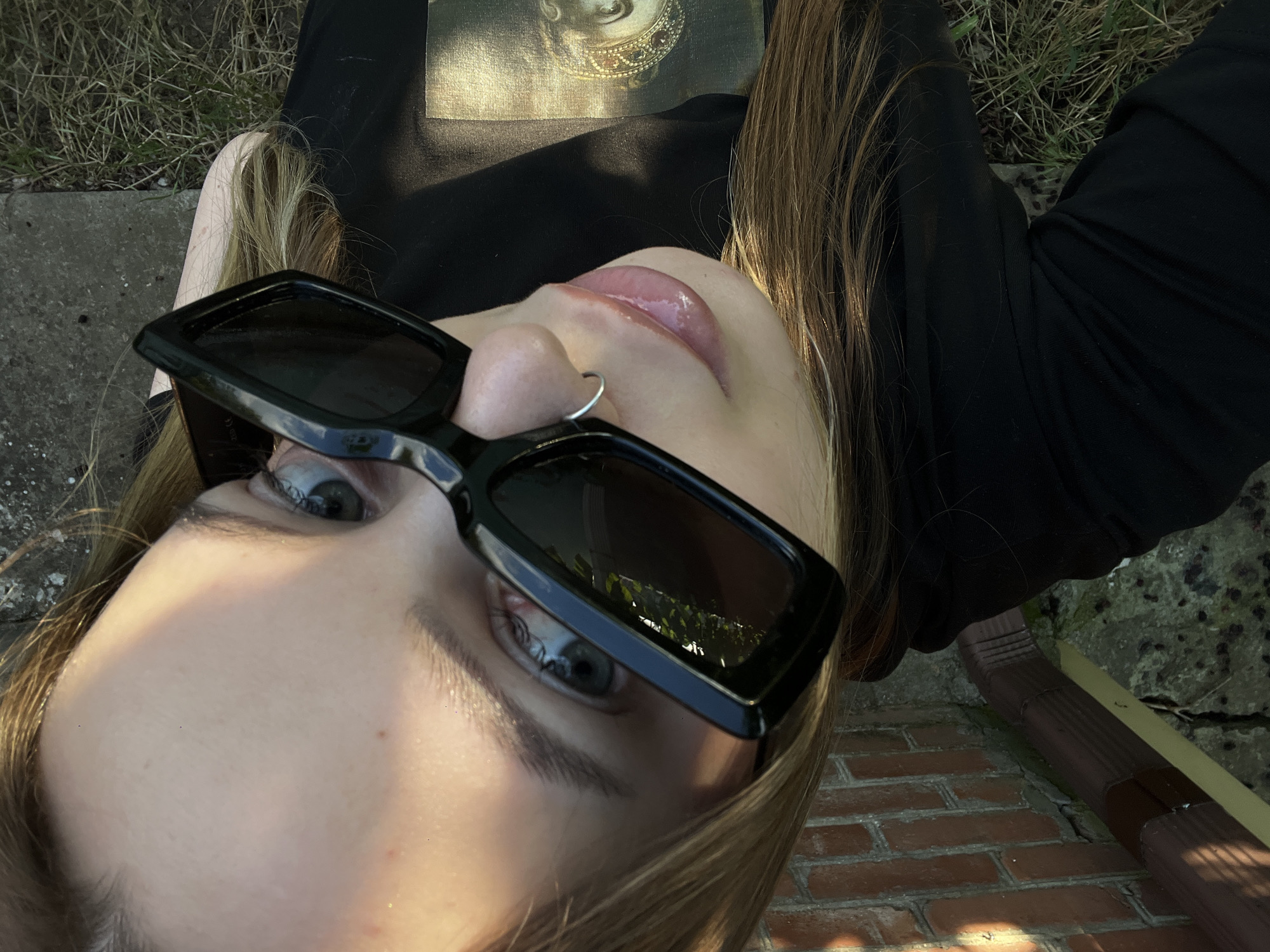 A close-up, overhead shot shows a young person lying down outdoors. They are wearing large, black, rectangular sunglasses that partially obscure their face. Their lips are slightly parted, and their tongue is visible. A small silver hoop piercing is visible on their nose. Their hair, a light brown, is spread out around them. They are wearing a black long-sleeved shirt with a faint image on it, possibly a portrait. The person appears to be relaxed.
The scene is outdoors with a mix of natural and man-made elements. In the background, there is dry grass and a rough, grey concrete edge. Below this, a textured red brick wall is visible, with a brown downspout running along it. The lighting suggests it is daytime, possibly late afternoon or early evening, with the sun casting shadows. The overall impression is casual and relaxed, suggesting the person is enjoying a moment outdoors. There is no visible text in the image.