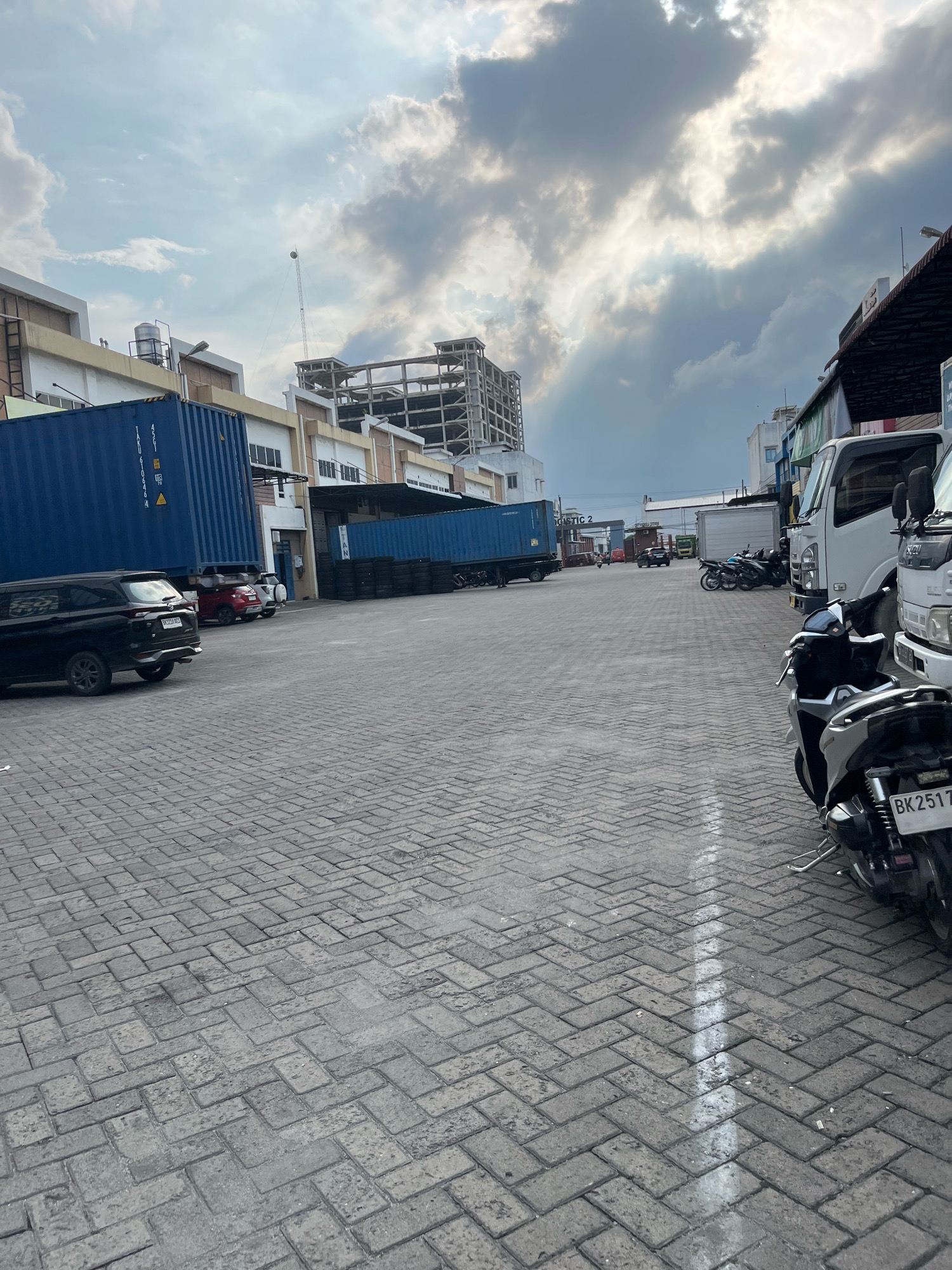 The image depicts an outdoor paved area, likely a logistics or industrial yard, in Percut Sei Tuan, Indonesia. The ground is covered with interlocking brick pavers arranged in a herringbone pattern. In the foreground on the right, a parked motorcycle is visible, with its license plate showing "BK 2517". Beyond the motorcycle, a white truck is parked.
To the left of the paved area, a black SUV is parked. Further back, several red cars are parked in front of a building with a blue shipping container. Another blue shipping container is visible further down the yard, positioned next to a stack of tires. In the background, there are several industrial buildings, including one under construction with visible scaffolding. The sky is partly cloudy, with sunlight breaking through the clouds. The overall impression is of a functional, working environment within an urban or industrial setting.