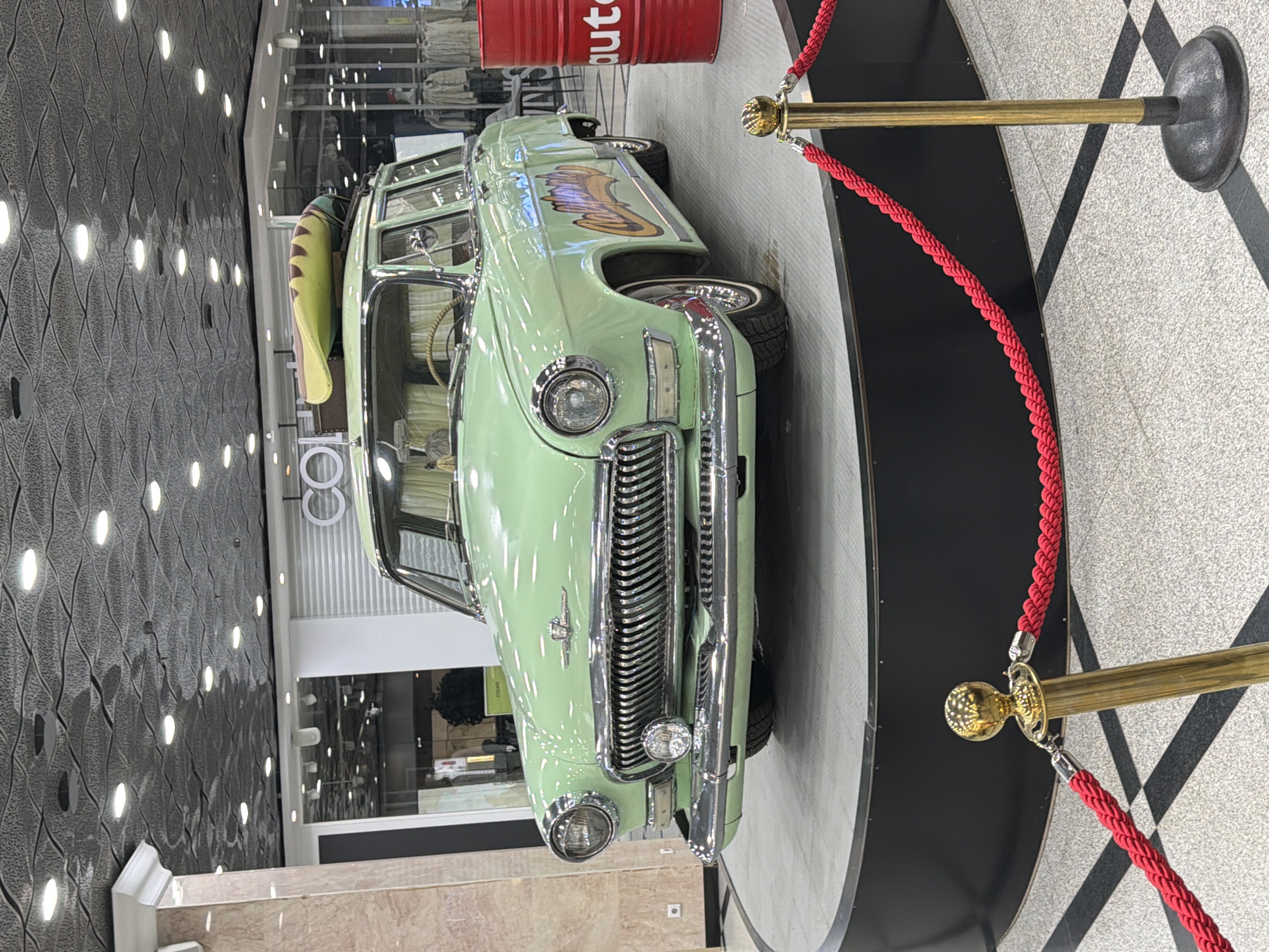 The image shows a light green vintage car on display, likely inside a shopping mall or exhibition space. The car features a chrome grill and a surfboard on the roof. A red barrel with "auto" written on it is visible behind the car. The car is surrounded by black and white tiled flooring and a red rope barrier attached to gold stanchions. The background suggests a modern interior with decorative lighting and a wall with a textured pattern. Considering the location provided, this could be a promotional display at a shopping center in Yekaterinburg, Russia, possibly advertising an automotive-related business or event.