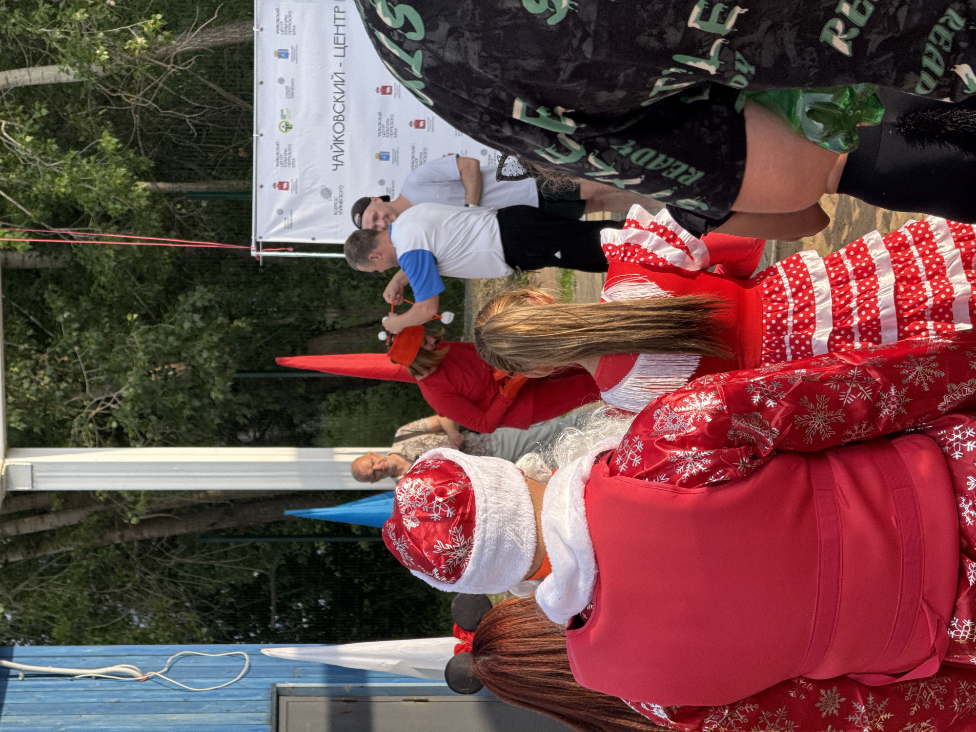 A group of people are gathered outdoors in front of a blue wooden building. In the foreground, a person in a red Santa Claus costume with a snowflake pattern is facing away from the camera. To their left, another person with auburn hair and black Mickey Mouse ears is also facing away. A young girl in a red and white polka-dot dress stands to the right of the Santa Claus figure. In the mid-ground, a man in a blue and white shirt and black shorts is interacting with a person in a light-colored t-shirt and black shorts. Behind them, a man in a light-colored shirt and grey pants stands looking towards the interaction. A white banner with Russian text is visible in the background. The environment appears to be a park or outdoor recreational area, with trees and foliage visible. The banner indicates the location is in Chaikovsky, Russia, specifically mentioning "Чайковский - центр", which translates to "Chaikovsky - Center". This suggests the event is taking place at a cultural or community center in Chaikovsky.