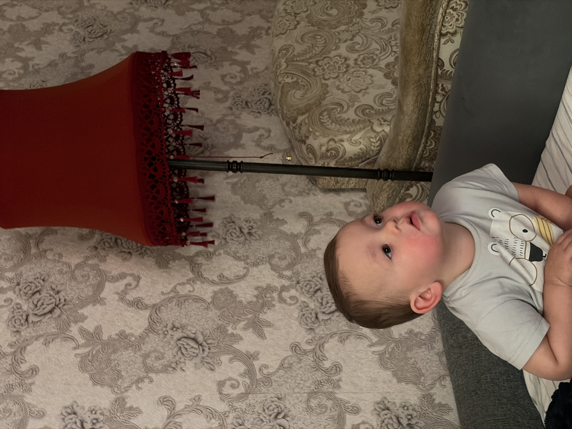 A light-skinned toddler sits on a gray sofa, looking to the right with a slightly open mouth. The toddler wears a light blue t-shirt with a cartoon bear graphic. The background features a textured wallpaper with a floral pattern and a red lampshade with fringed trim. To the right of the toddler is an ornate sofa with a similar floral pattern. This image was captured indoors.